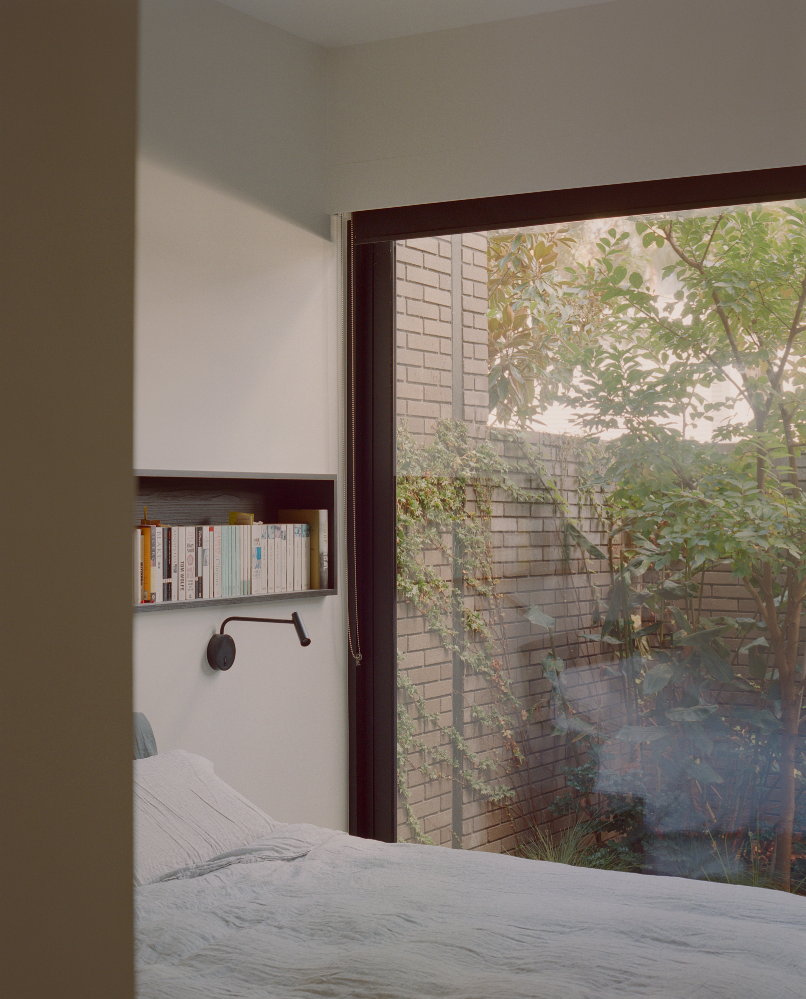 Bedroom corner with recessed shelf and full-height window overlooking a brick courtyard wall with tree