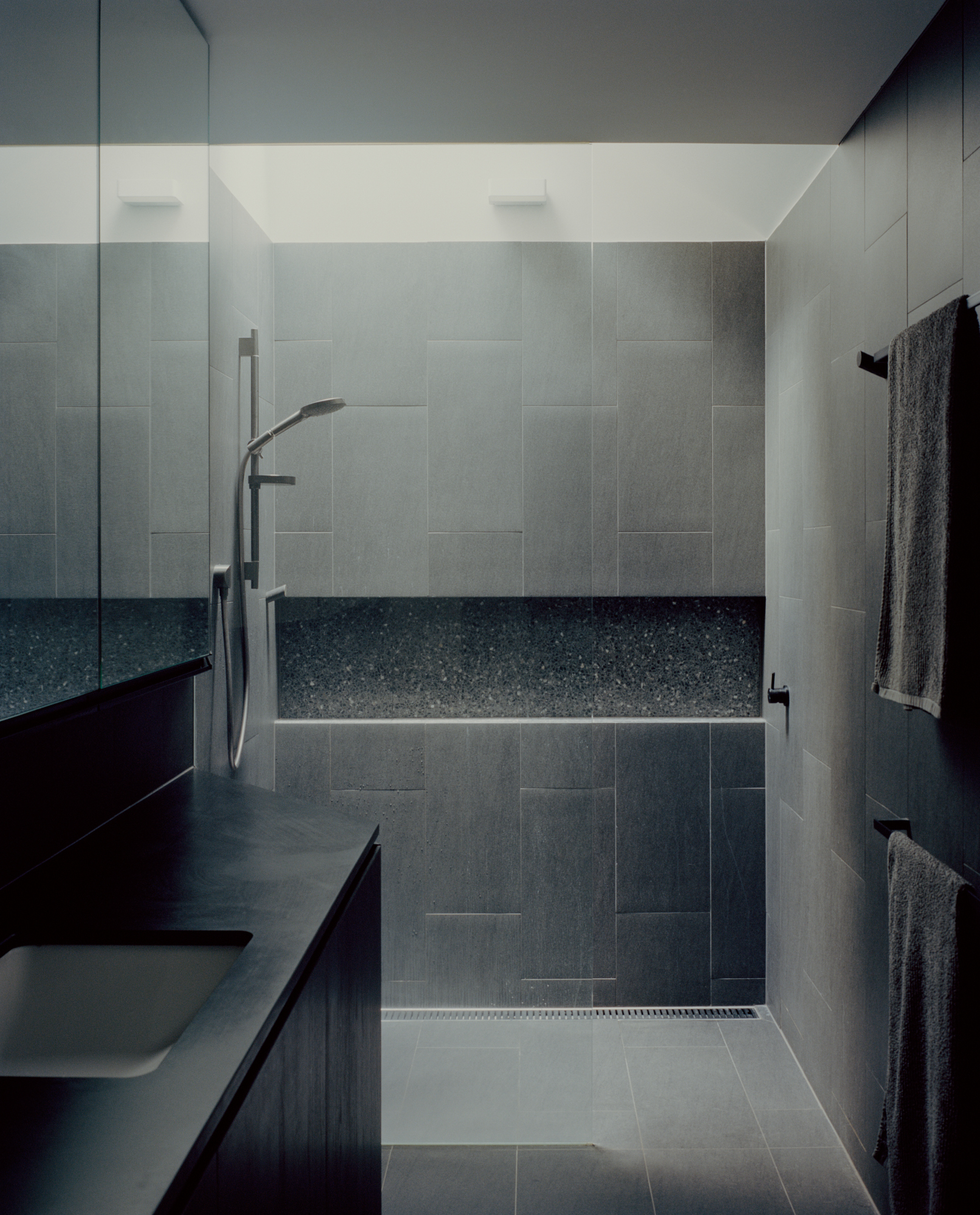 Walk-in shower enclosure with large-format grey tile walls and terrazzo bench under recessed ceiling lights