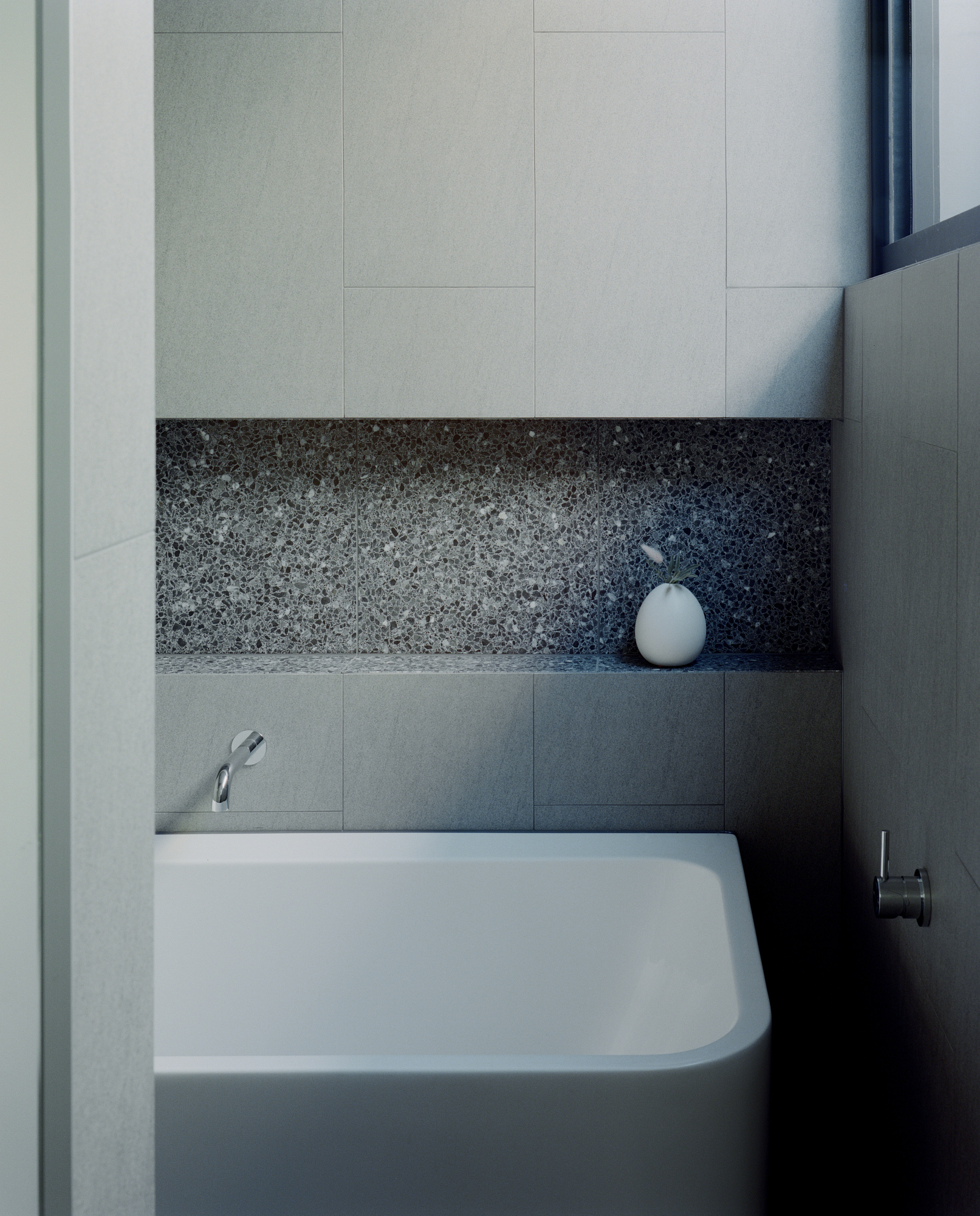 Freestanding white bathtub against terrazzo niche with grey tile surround and wall-mounted faucet