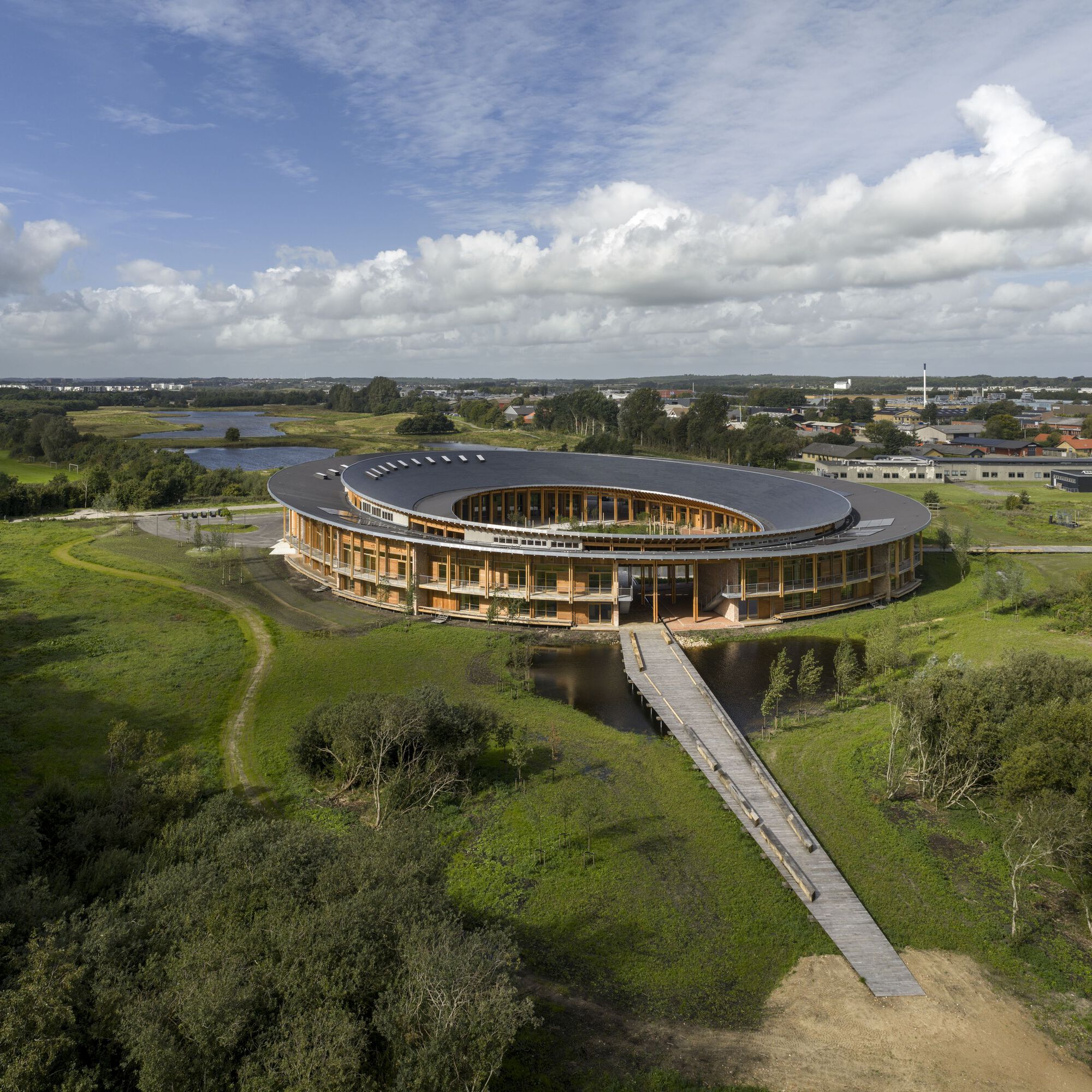 Aerial view of the ring-shaped structure with dark roof and timber colonnade approached by a wooden boardwalk