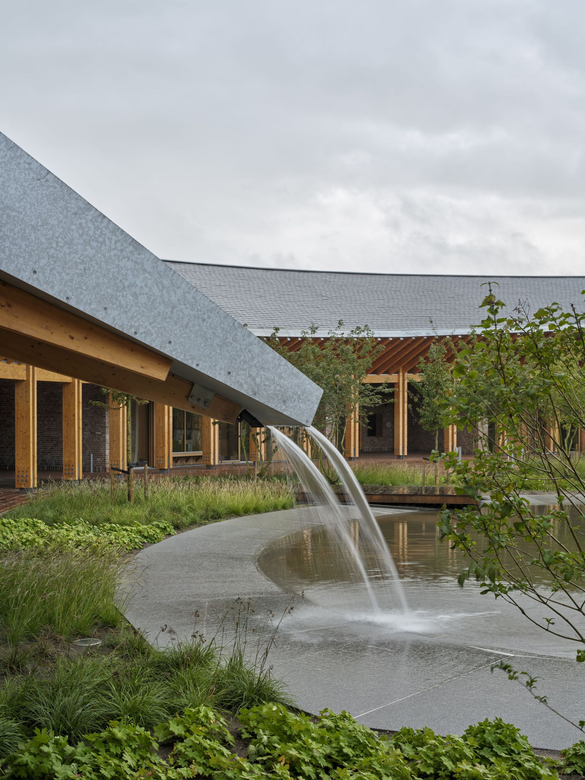 Courtyard water fountain with curved spout beside planted beds and overcast sky