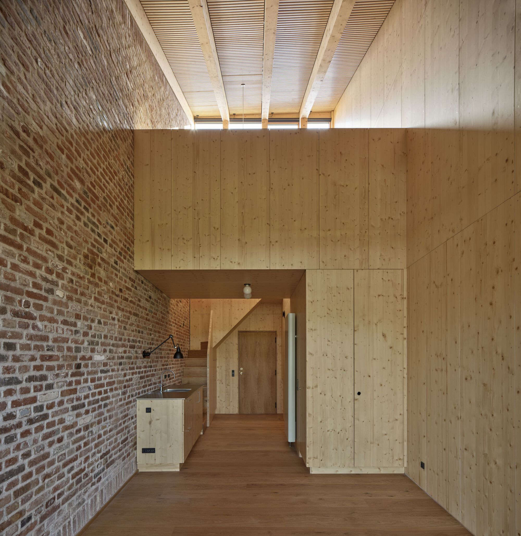 Double-height room with exposed brick wall, plywood volume, and slatted timber ceiling with clerestory windows