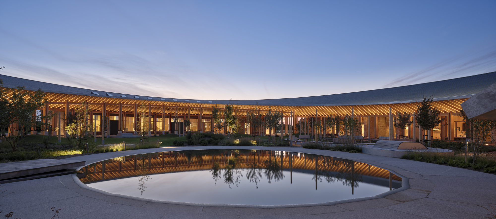 Circular reflecting pool with curved timber colonnade illuminated at twilight