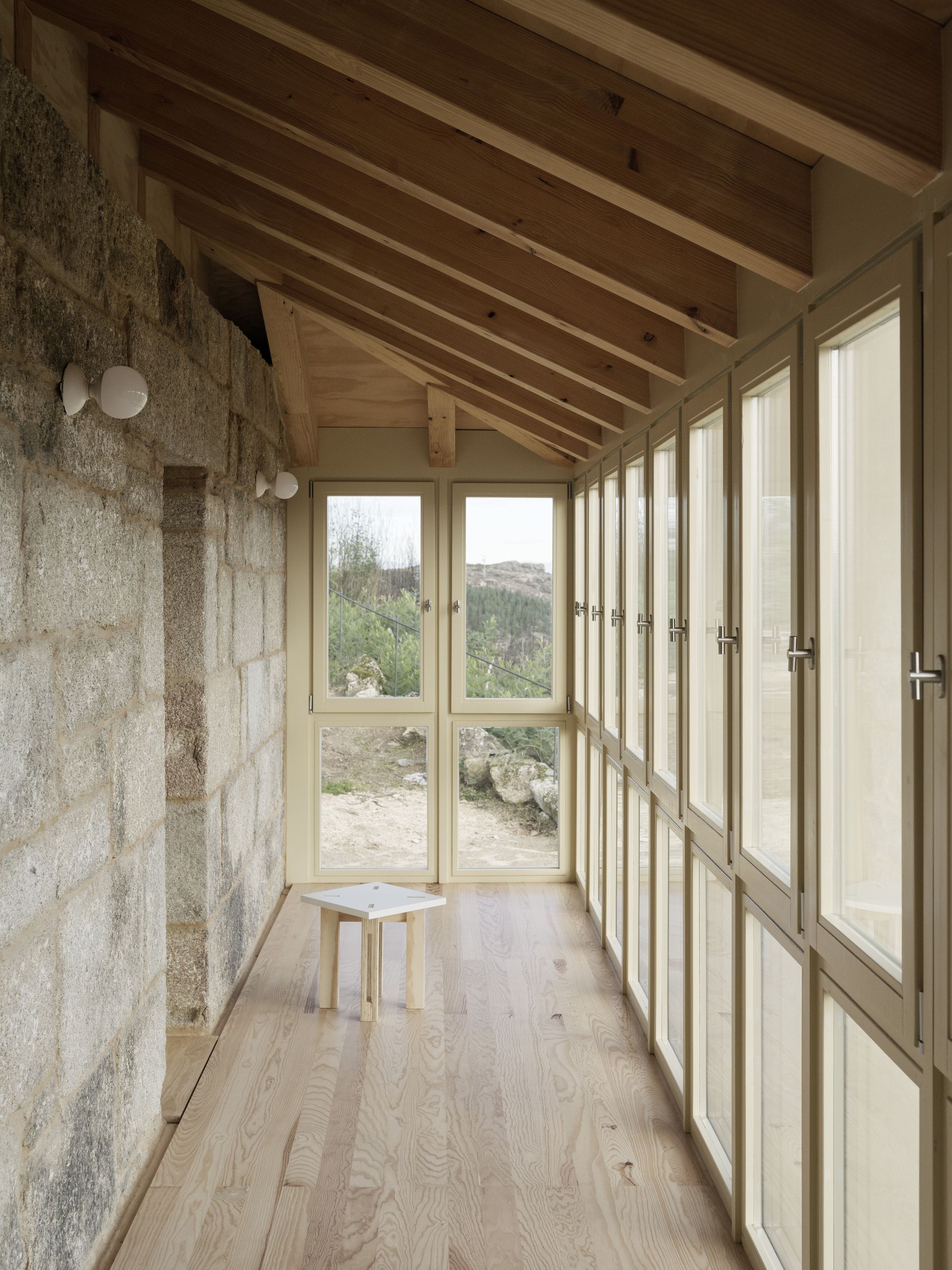 Narrow glazed corridor with timber floor, exposed rafters and stone wall overlooking the landscape