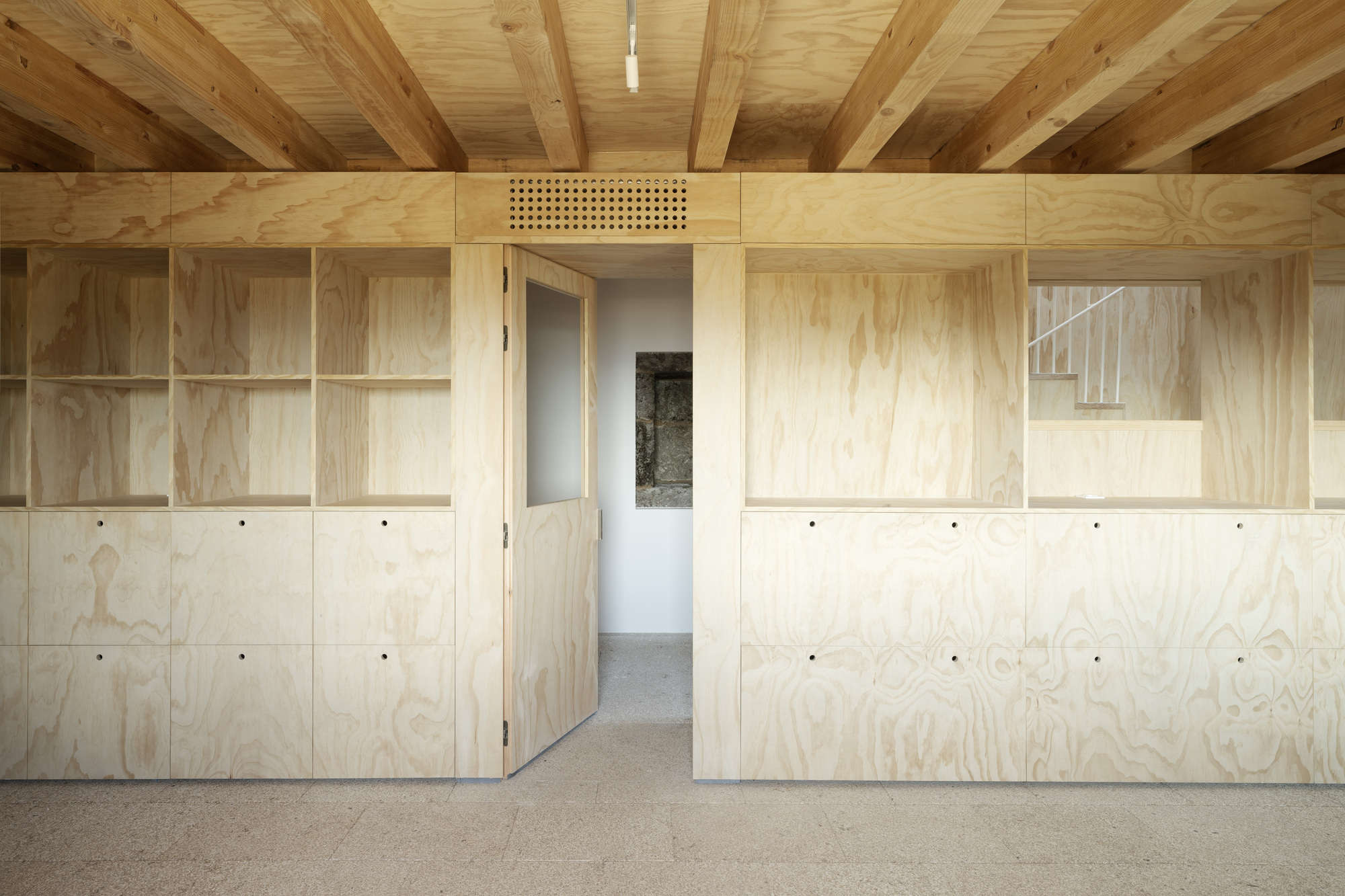 Interior wall of plywood cabinets and shelving with exposed timber beams overhead and a perforated ventilation panel