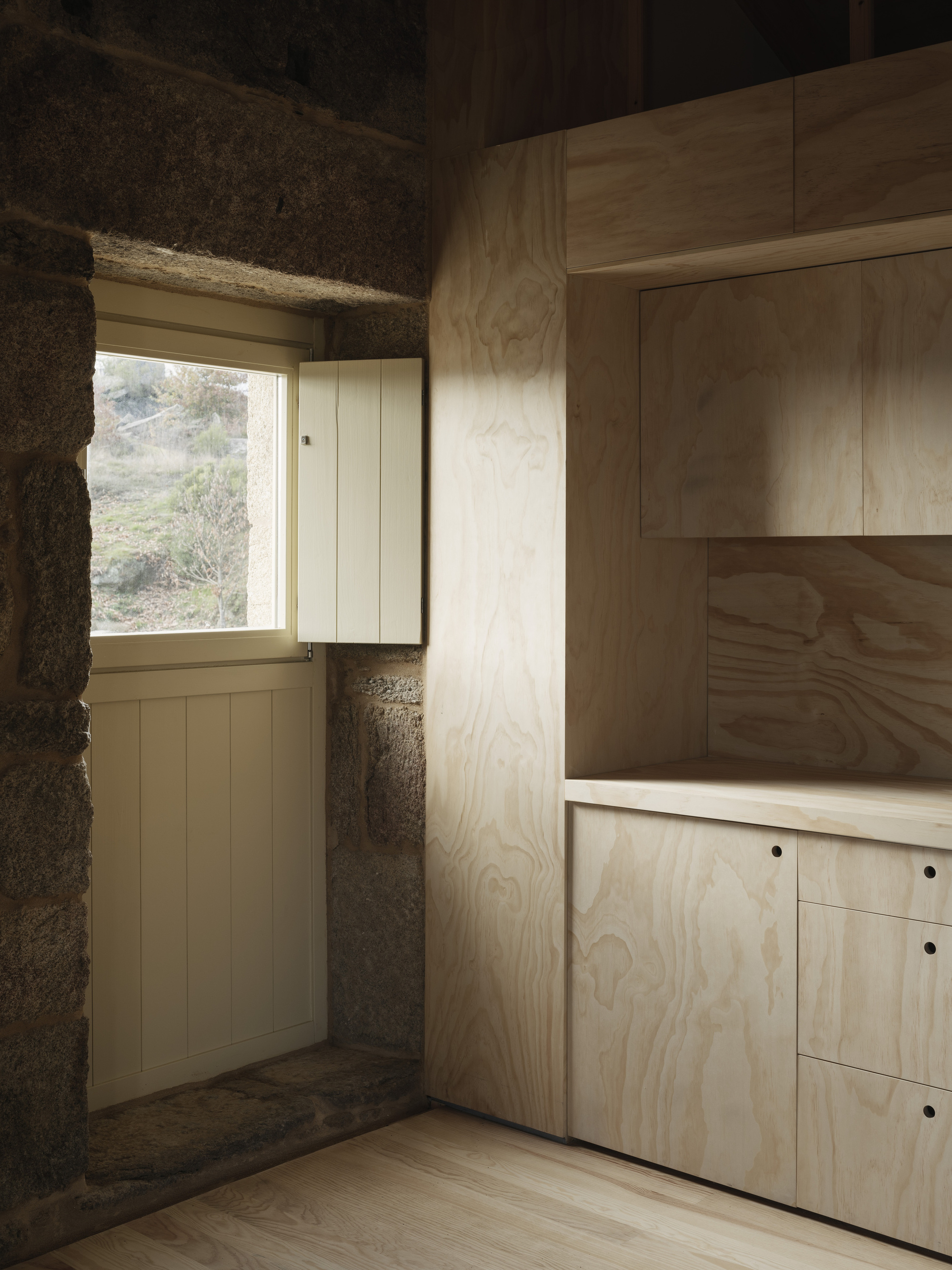 Corner window framed by exposed stone walls and plywood cabinetry with afternoon light streaming through