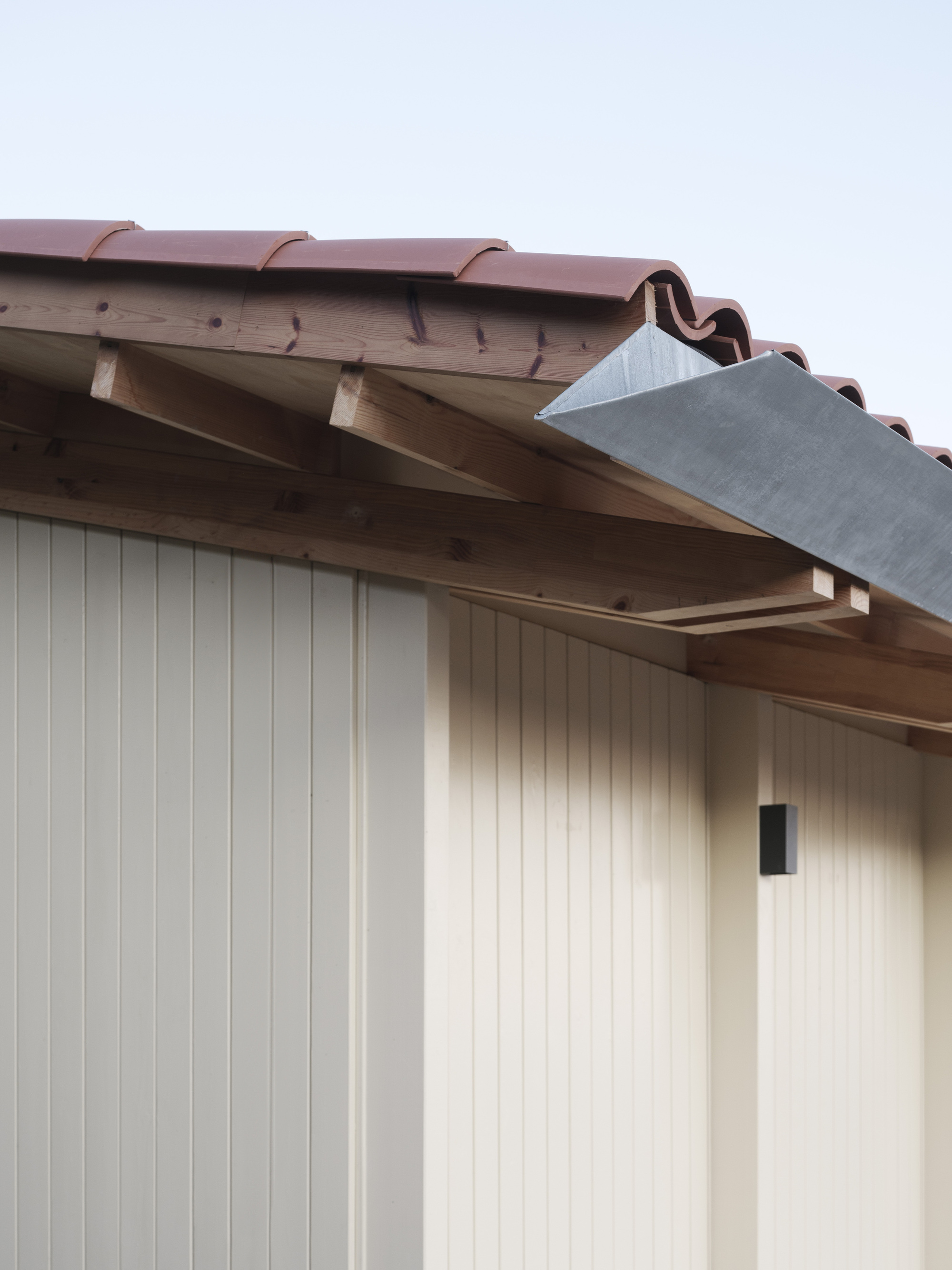 Eave detail showing timber rafters, terracotta roof tiles, and vertical paneled cladding against an overcast sky