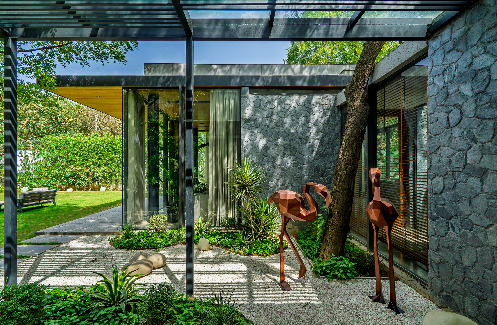 Steel pergola framing a view of the tree trunk wrapped in stone wall with gravel and planted ground cover