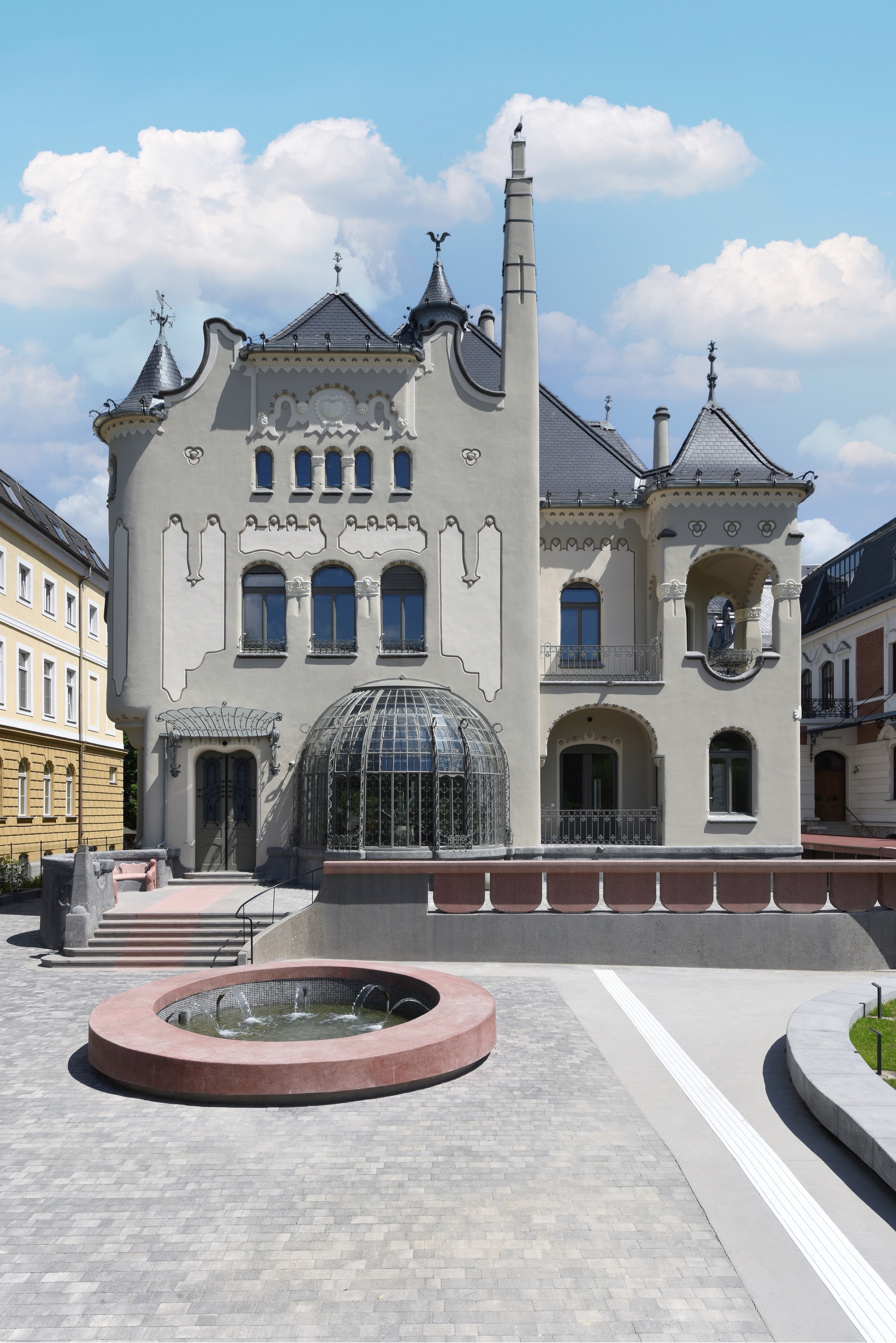 White stucco facade with domed glass pavilion, turrets and circular fountain in a public square