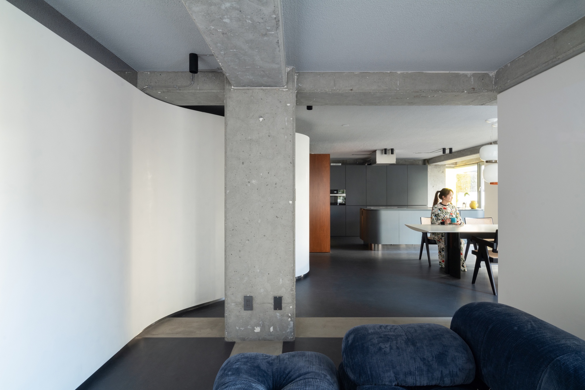 View through exposed concrete column toward open kitchen with person standing at counter in afternoon light