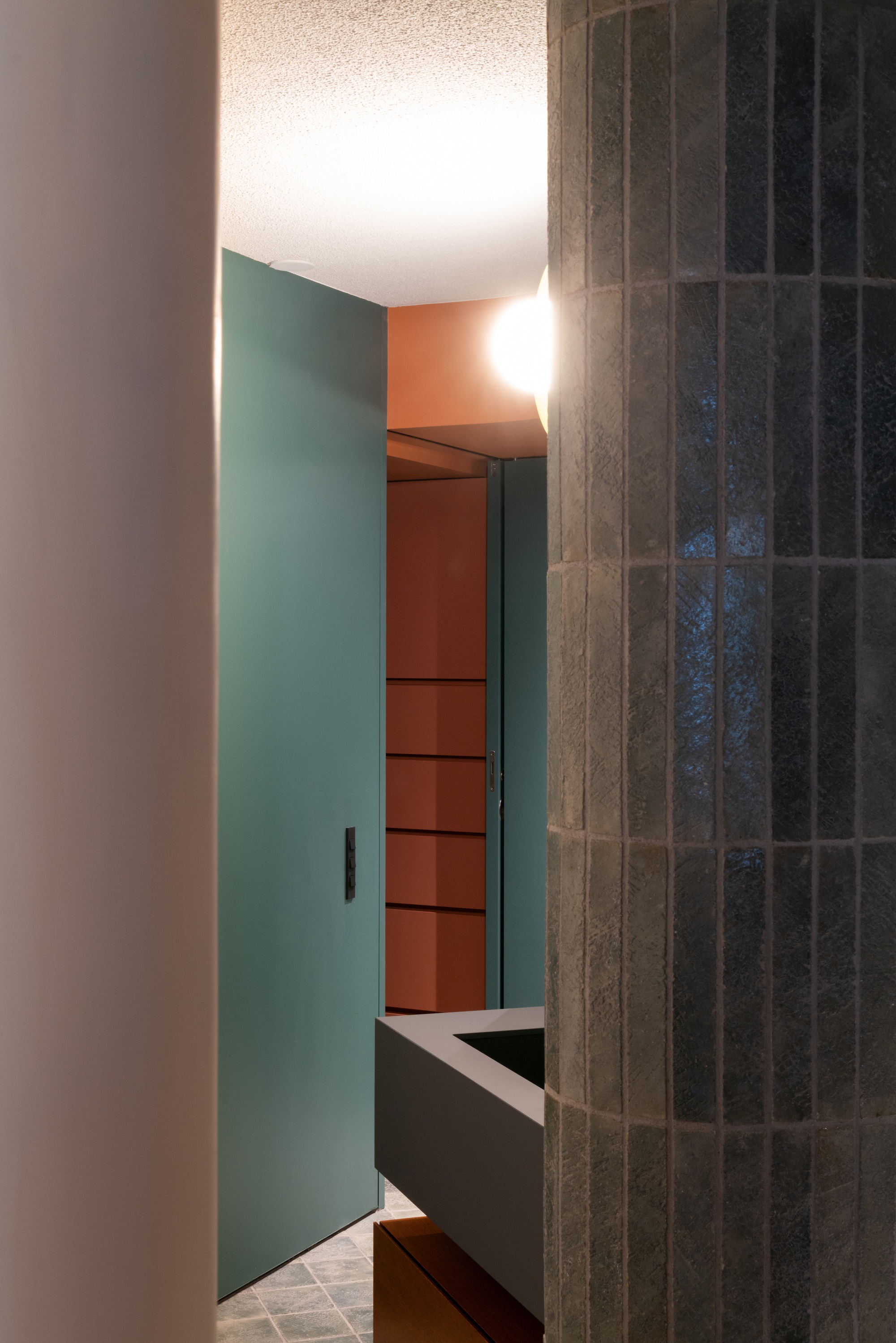 Doorway framed by teal wall panels and dark vertical tile leading to terracotta shelving beyond