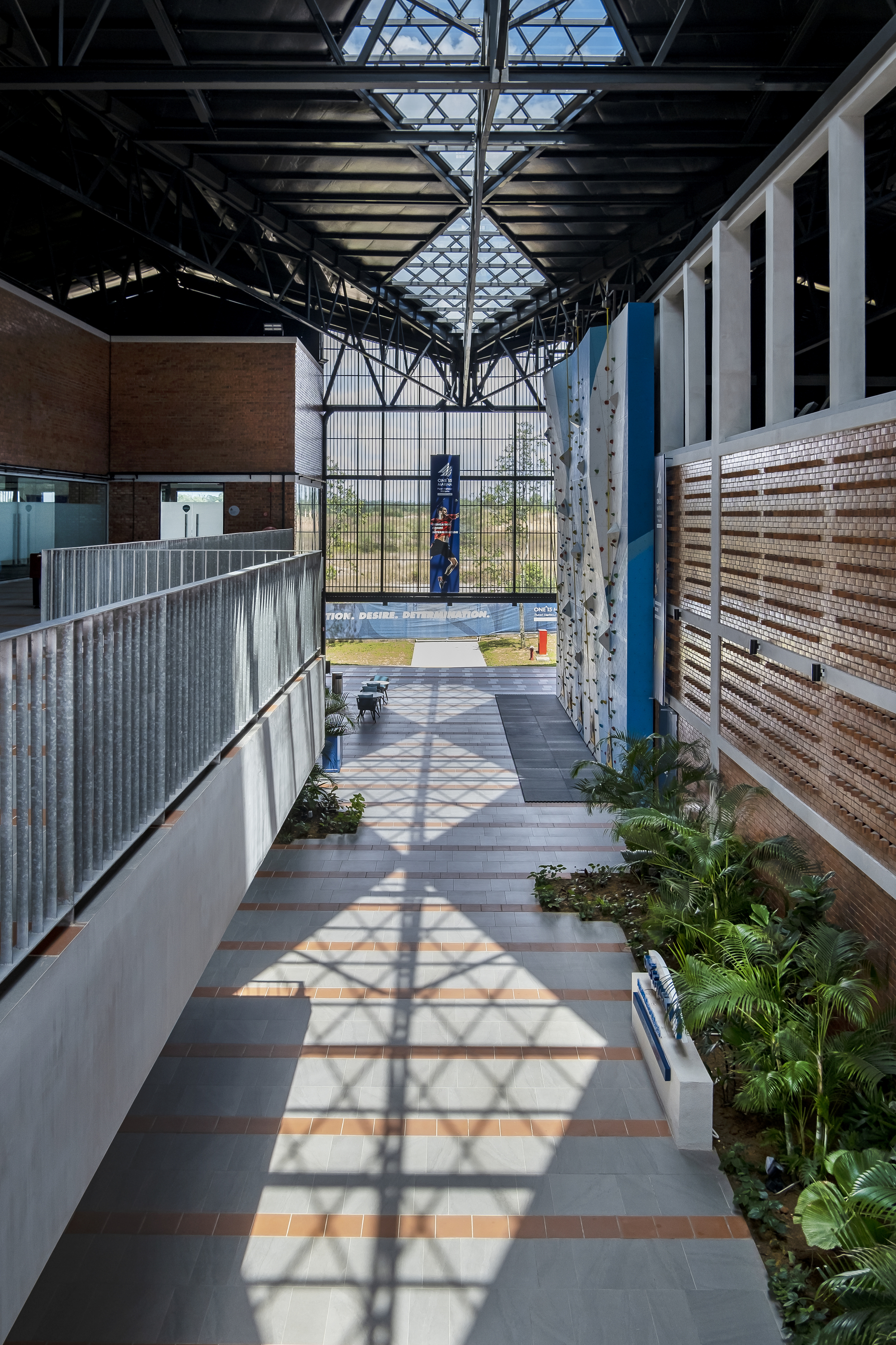 Interior corridor with exposed steel trusses, patterned shadows, planted beds, and glazed end wall