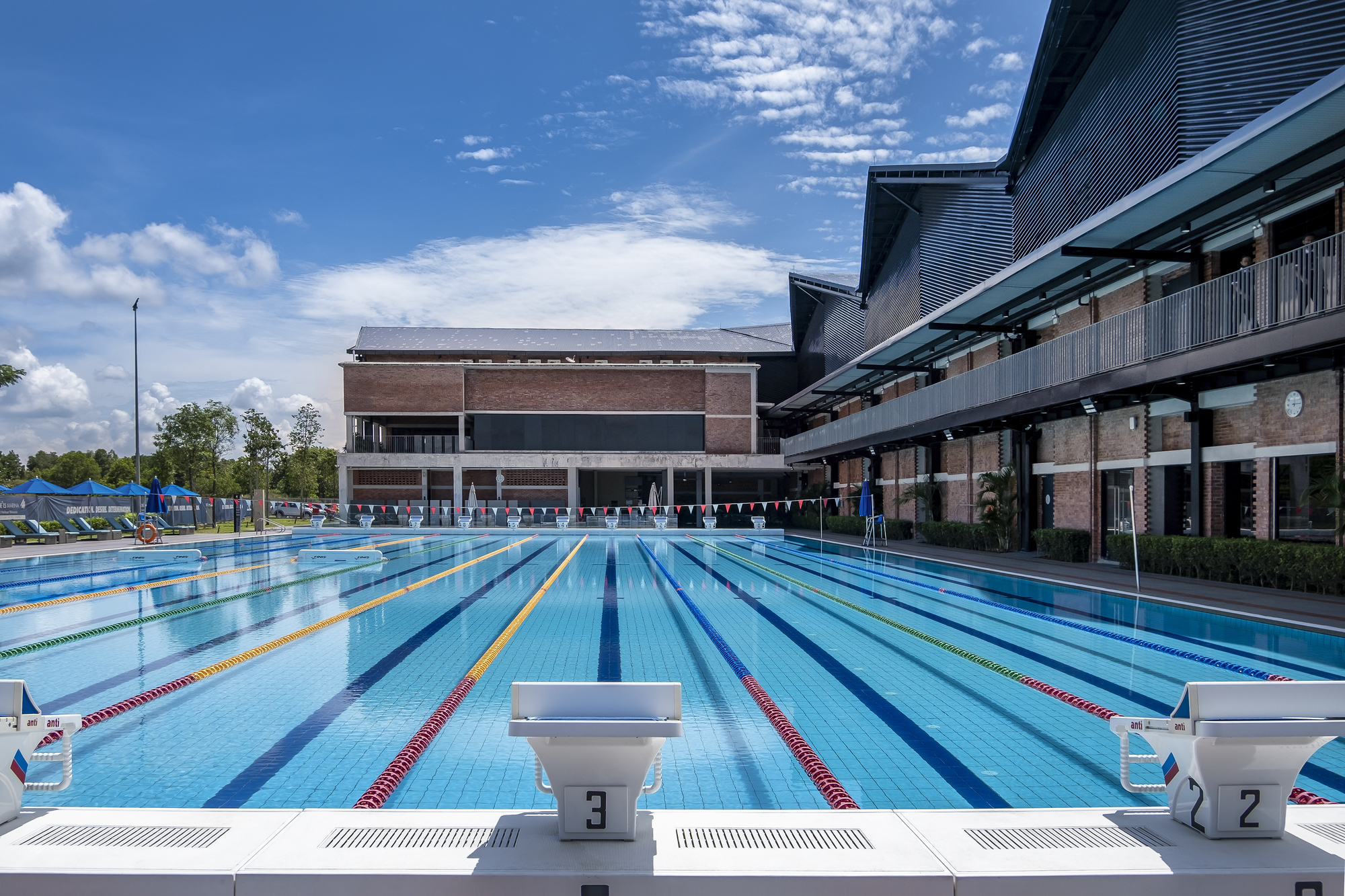 Olympic-sized swimming pool with lane markers, starting blocks, and brick pavilion under scattered clouds