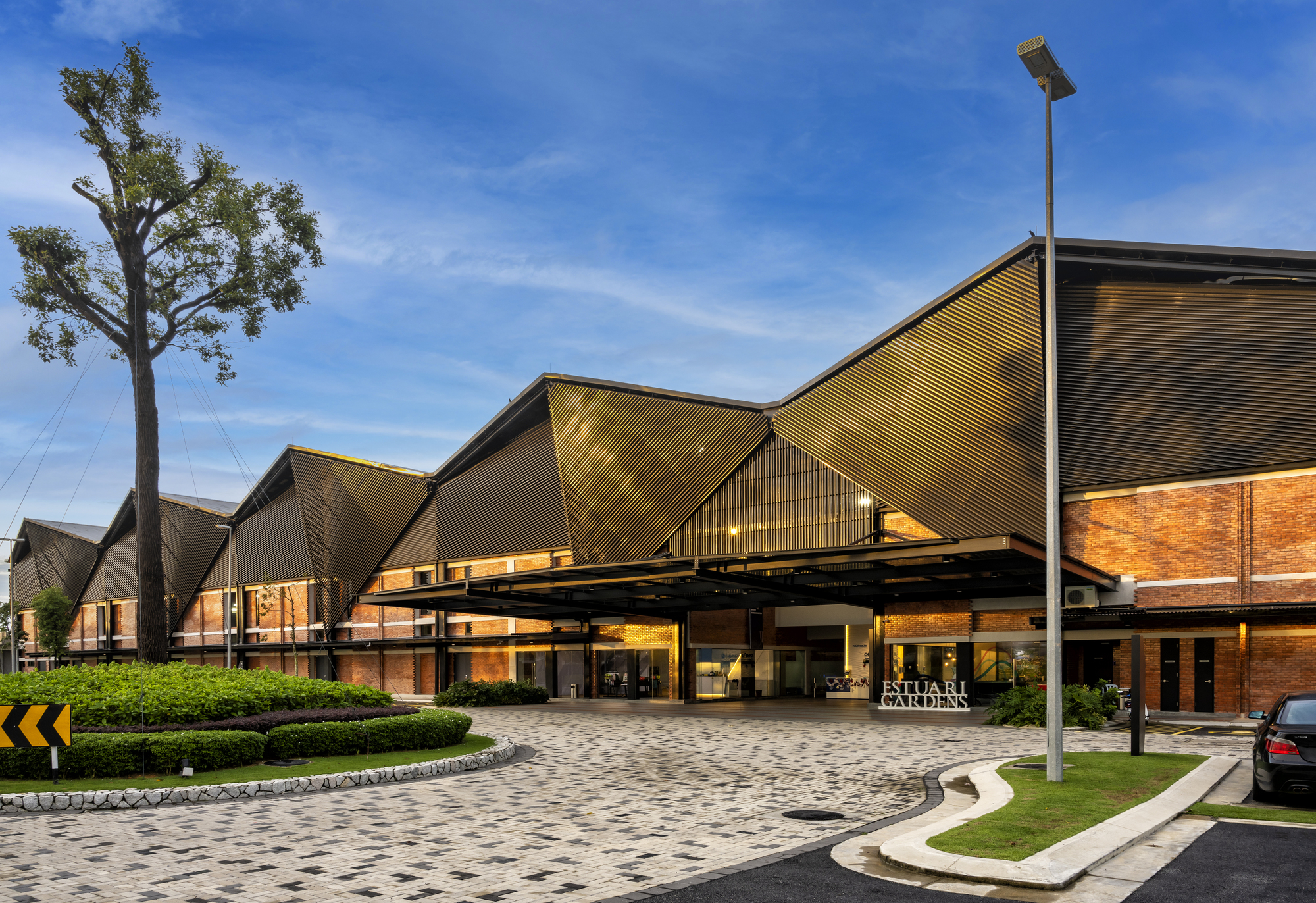 Entry canopy with folded metal roof planes over a paved forecourt with mature trees