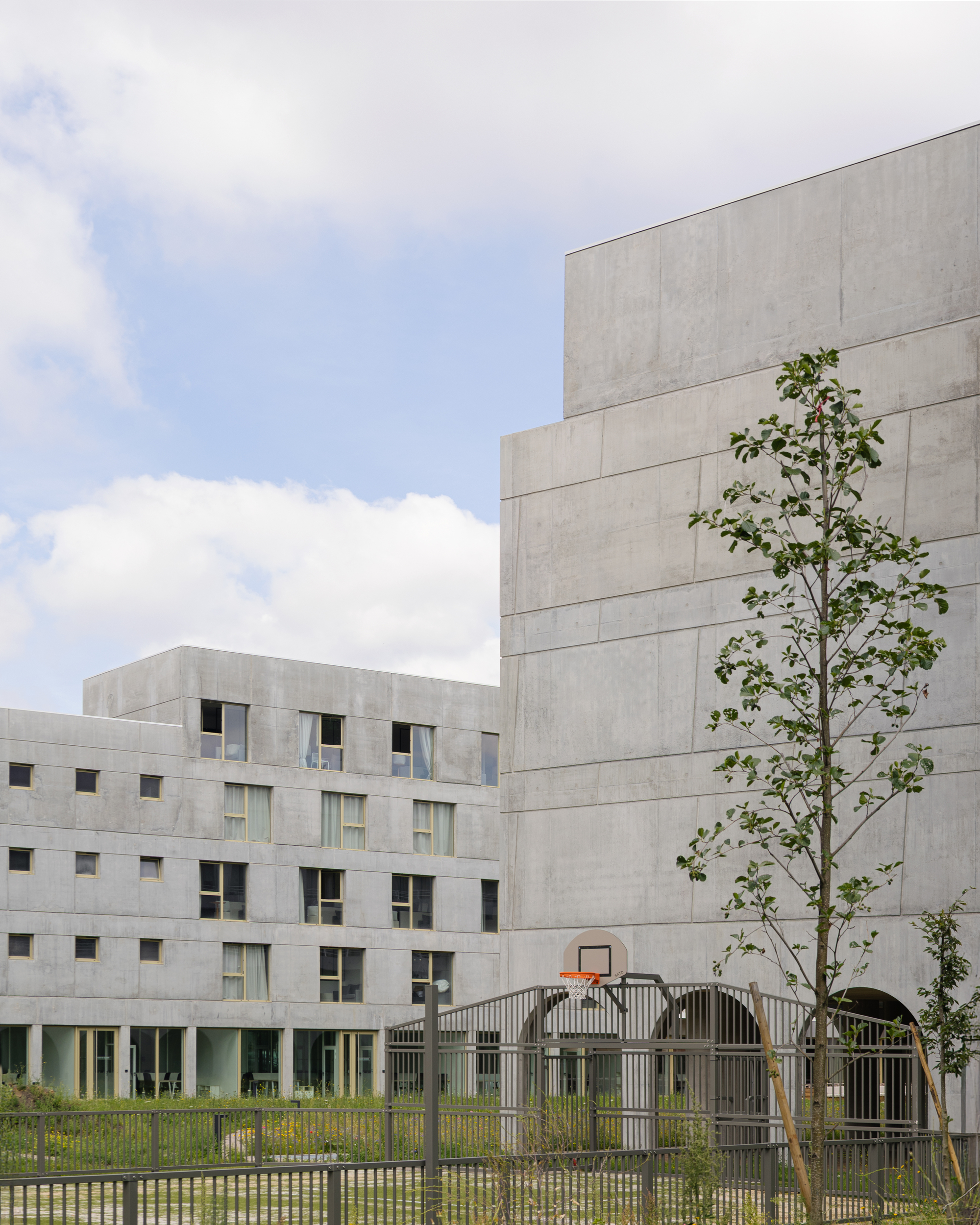 Concrete block facade with small punched windows beside a young tree and metal fenced play area