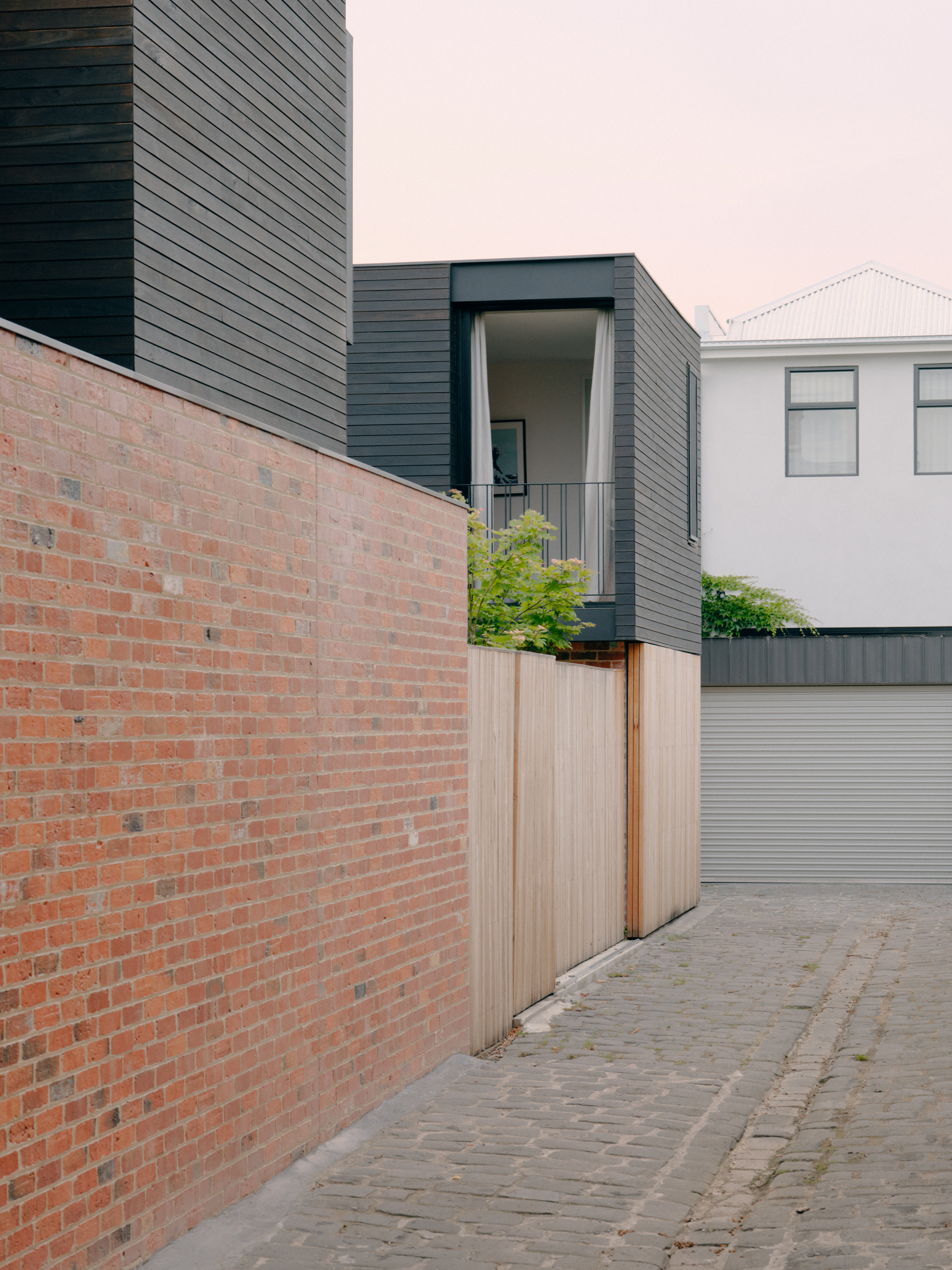 Narrow laneway with red brick boundary wall and dark horizontal cladding on upper volumes under overcast sky