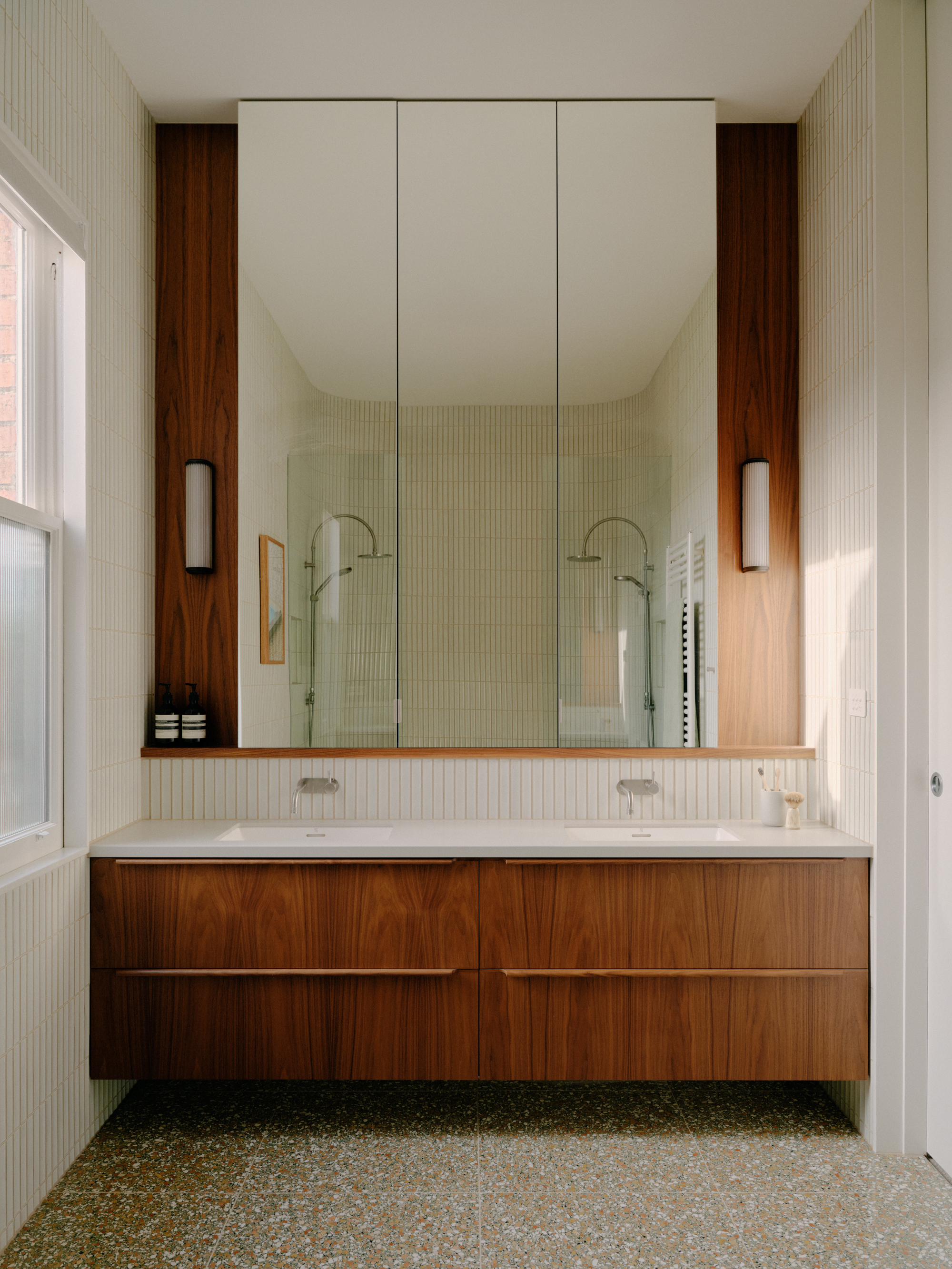 Floating walnut vanity with dual sinks and full-height mirrored cabinet in a tiled bathroom
