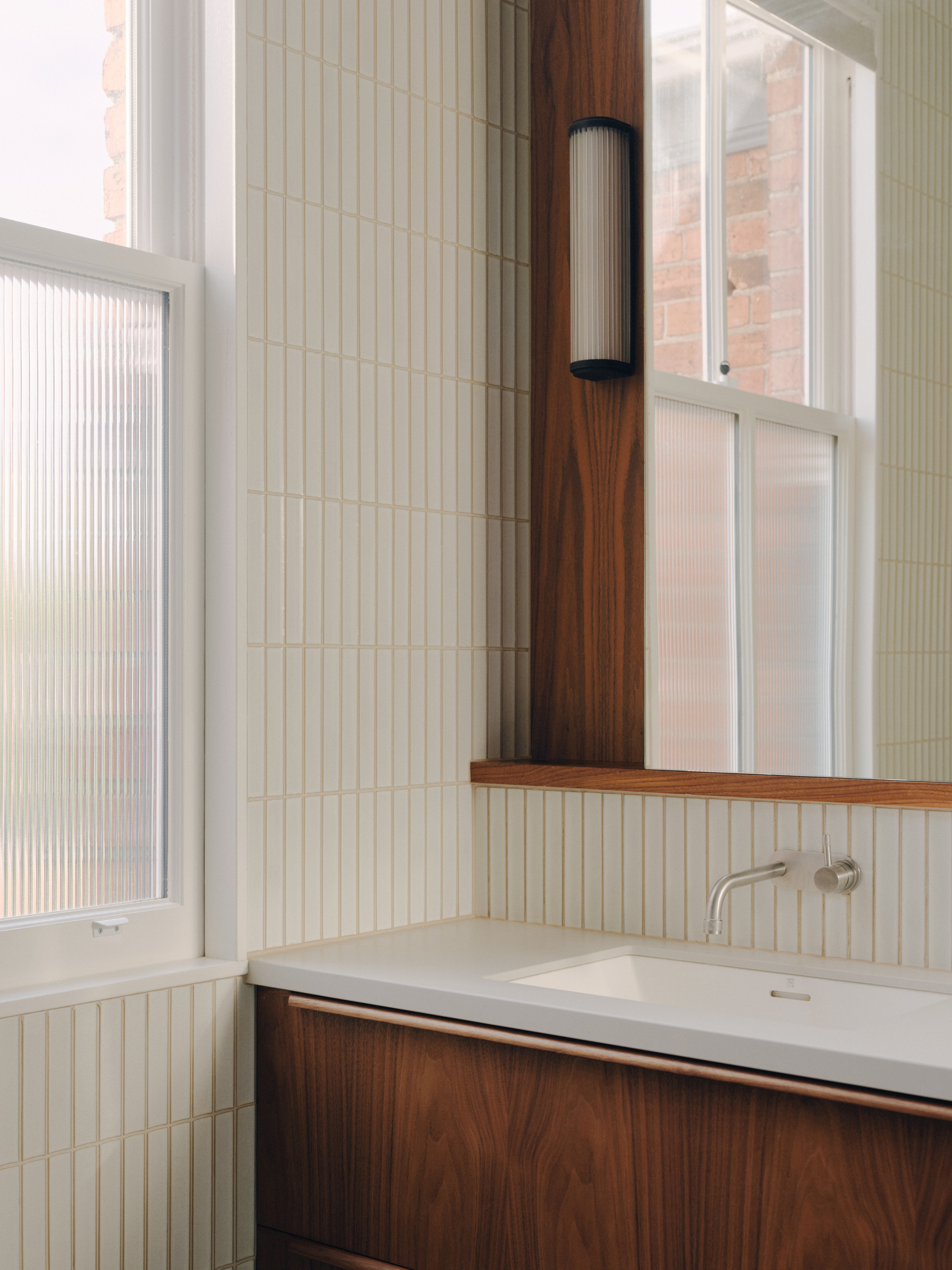Bathroom vanity corner showing vertical tile, walnut shelf, and cylindrical wall sconce near window