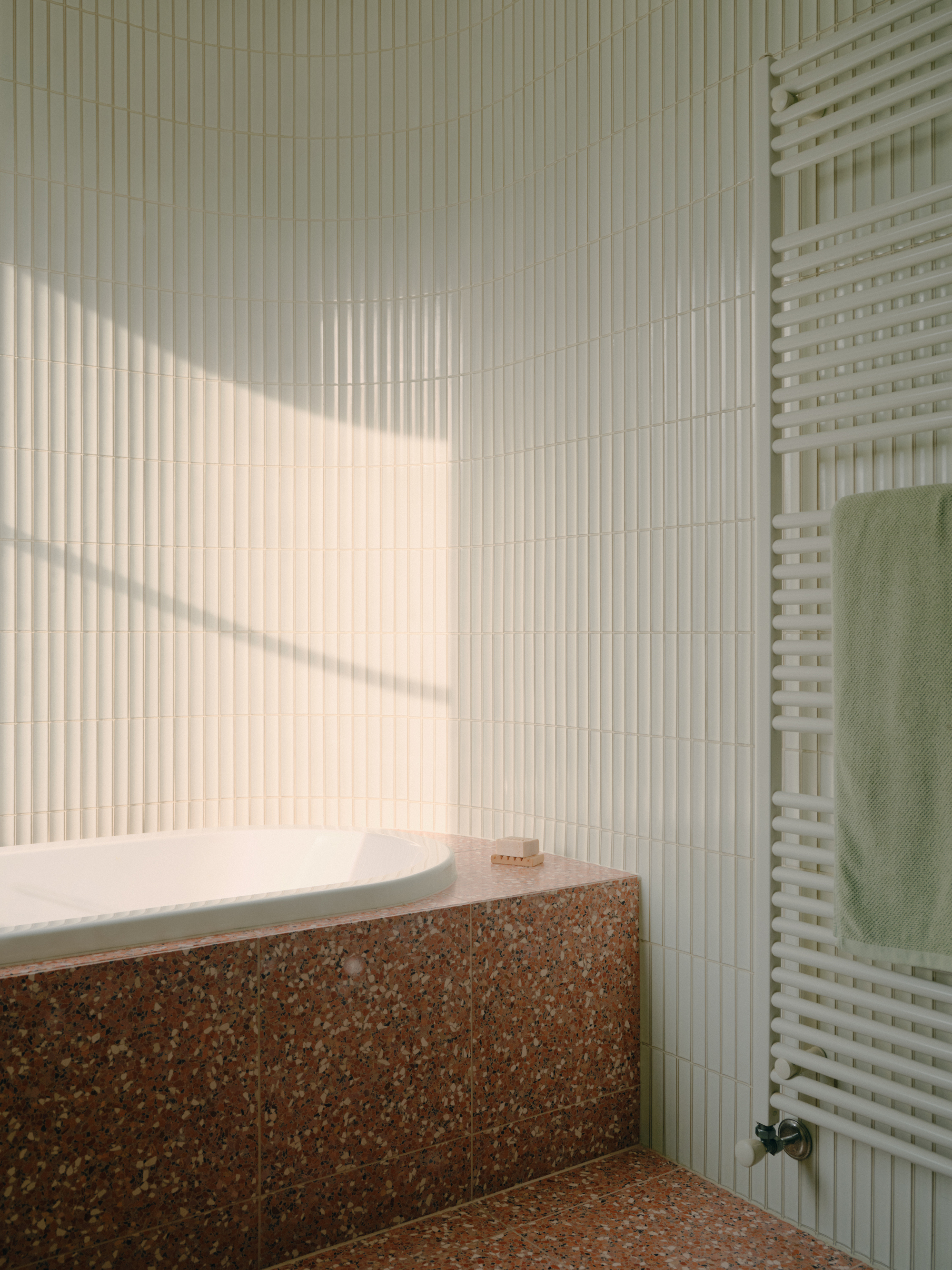 Bathtub alcove with terrazzo cladding and vertical ceramic tile catching afternoon sunlight