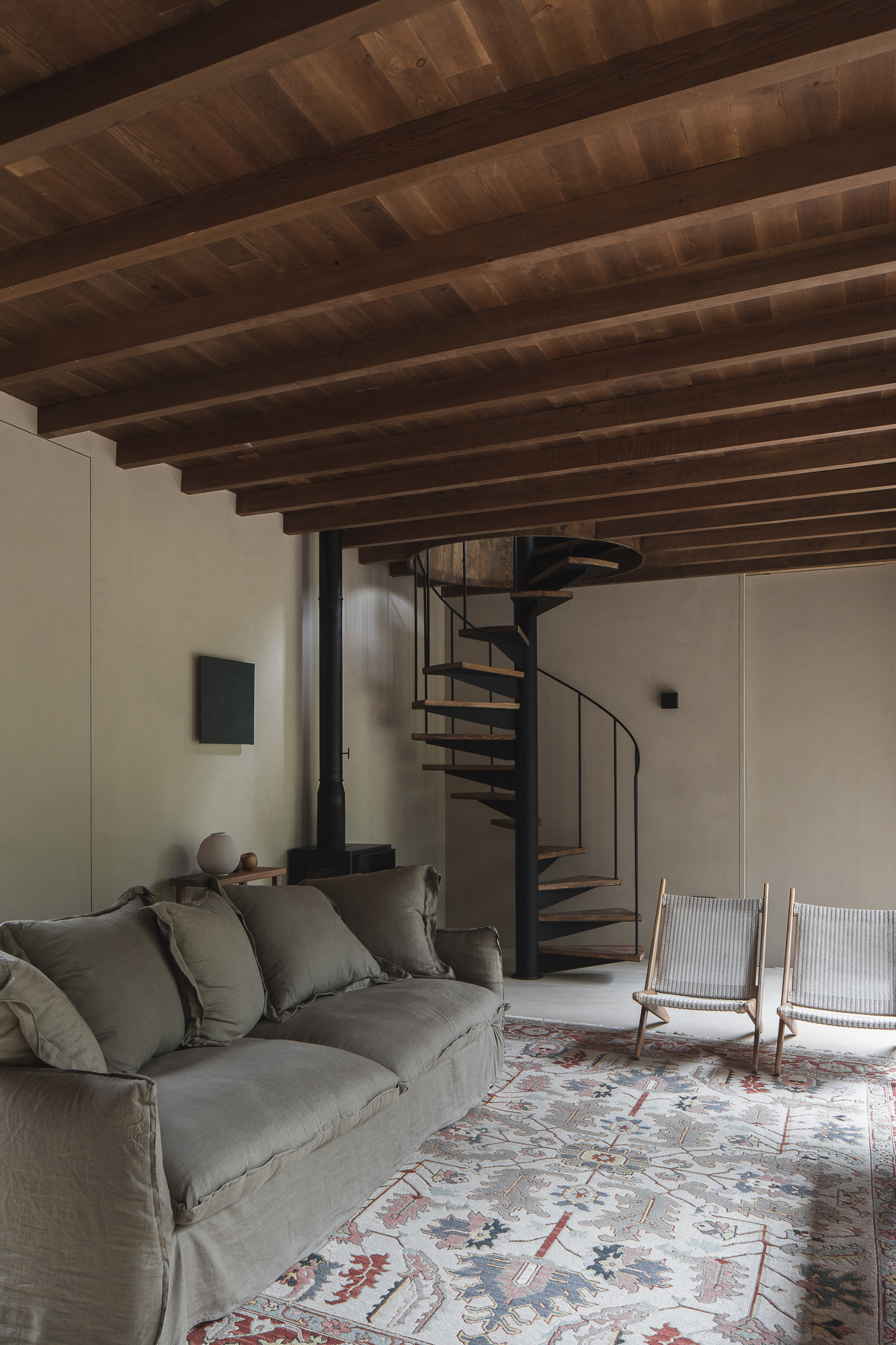 Living room with exposed timber beam ceiling, steel spiral stair, and patterned rug under diffused natural light