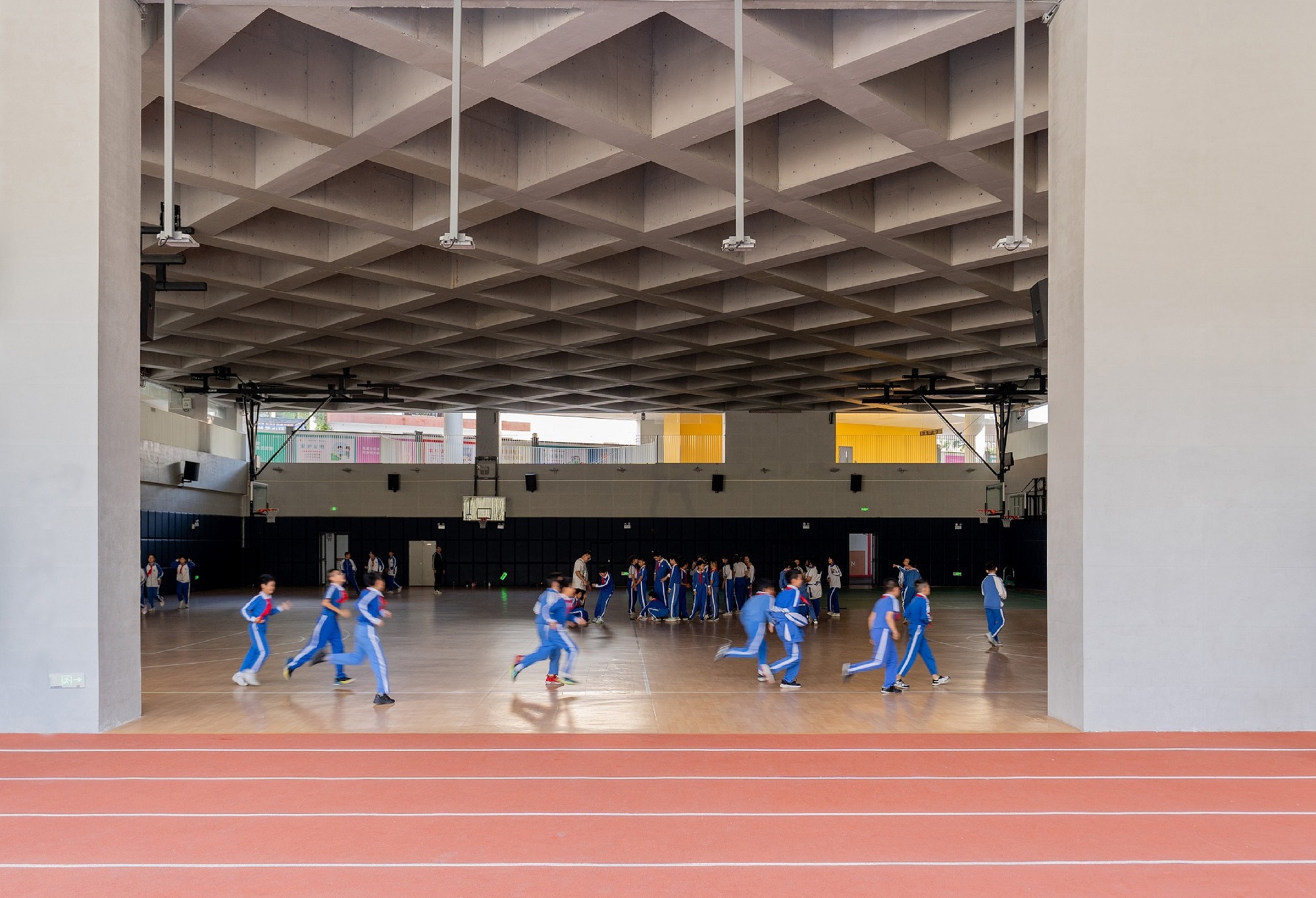 Indoor gymnasium with coffered concrete ceiling and students playing basketball on wood floor