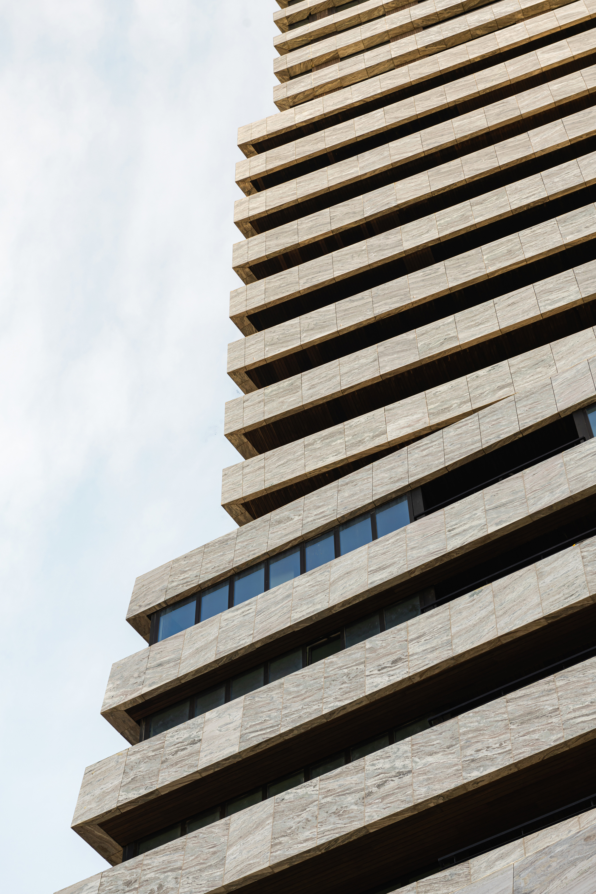 Upward view of stacked balconies with deep soffits and horizontal stone cladding against cloudy sky