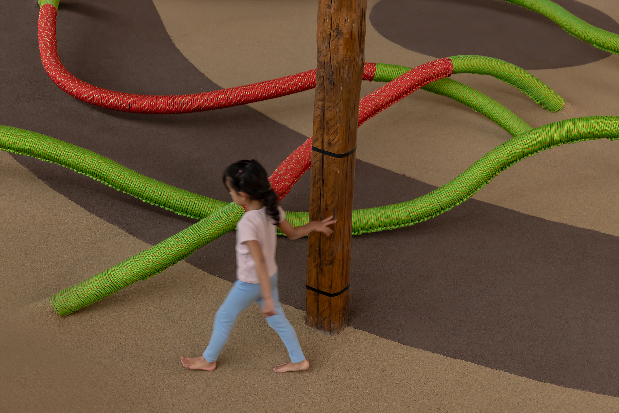 Child walking past timber column with green and red rope tubes extending across floor