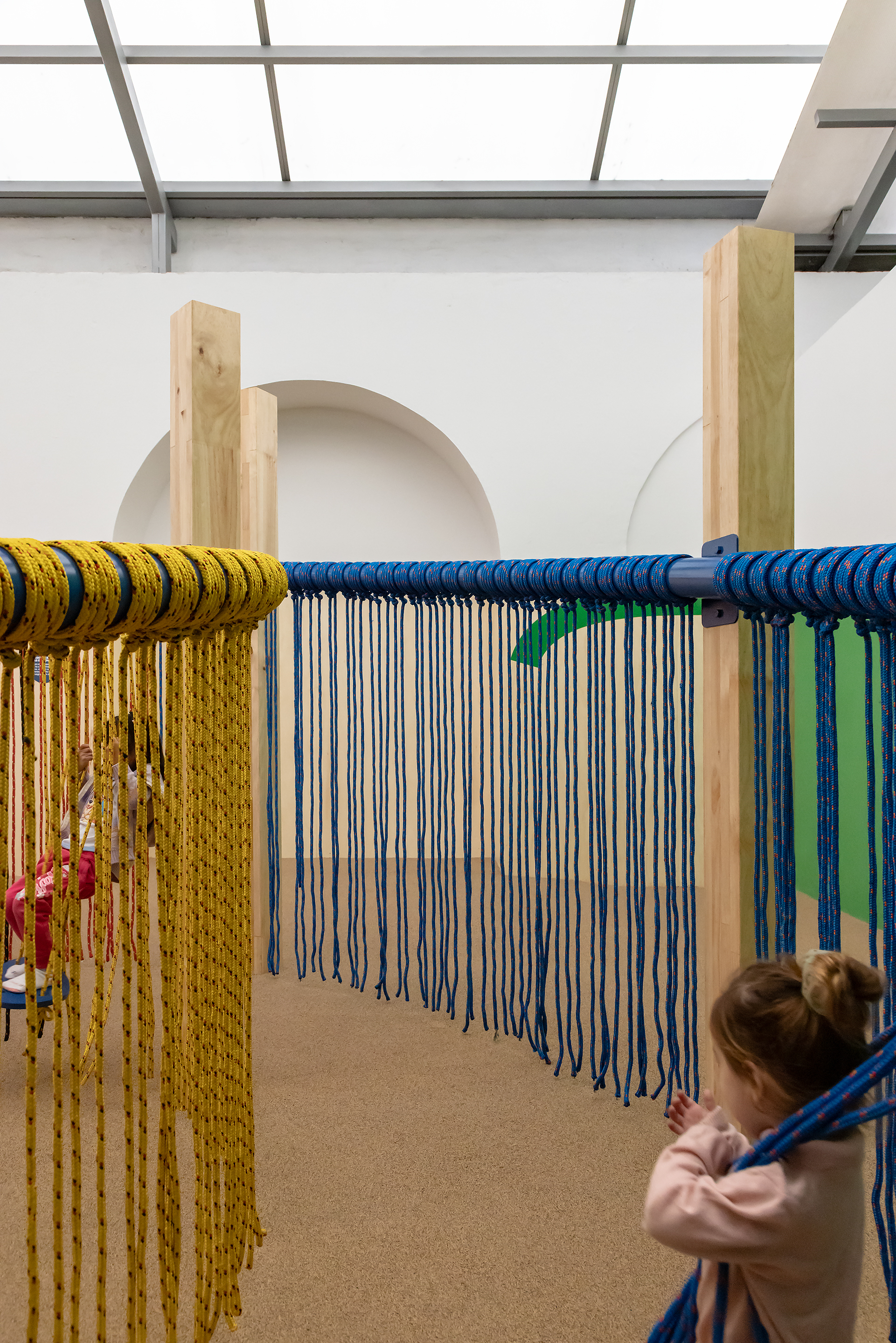 Interior installation with timber frames and yellow and blue ropes hanging vertically as a child reaches out