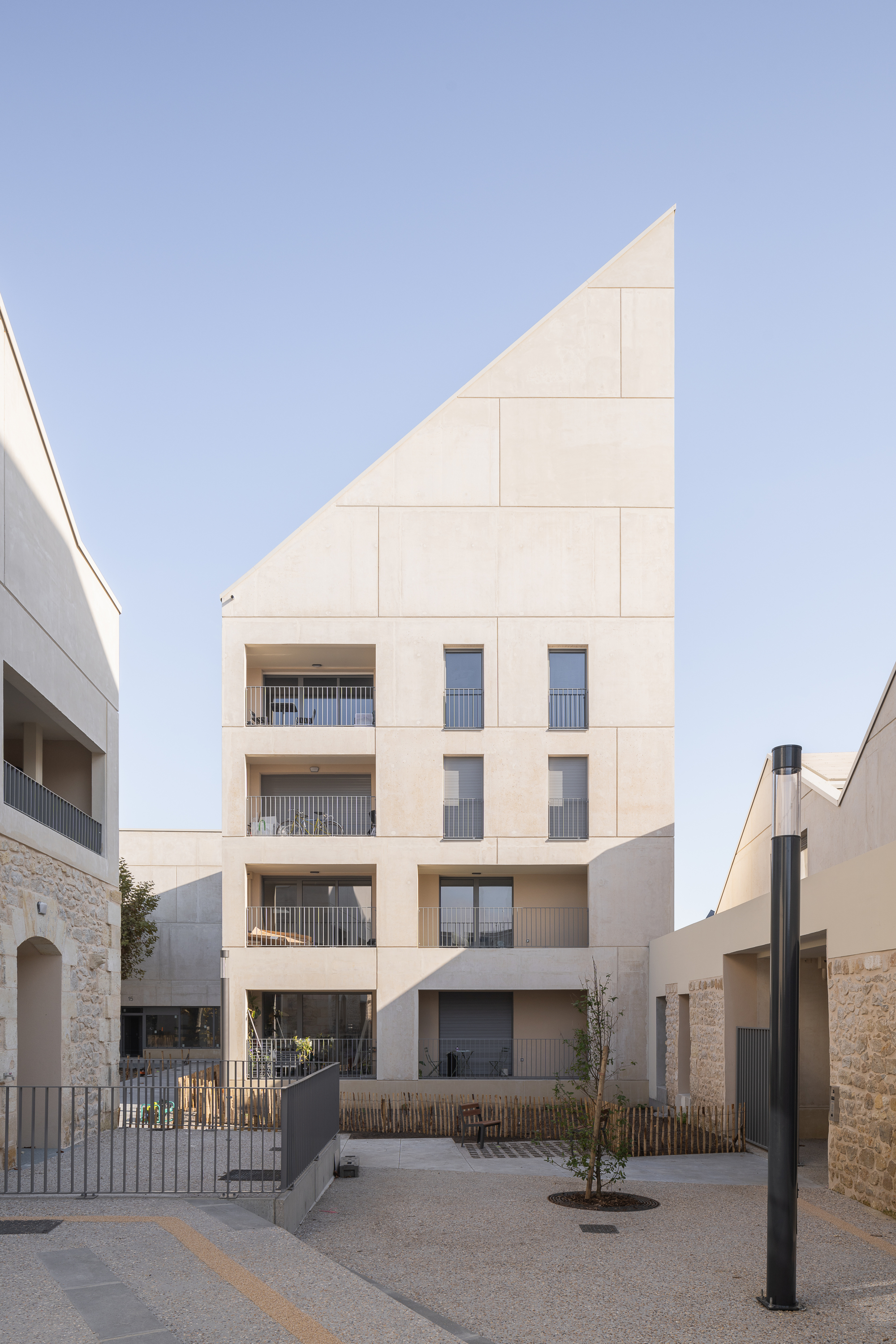 Angled facade with diagonal roofline clad in pale stone panels and recessed balconies facing a gravel courtyard