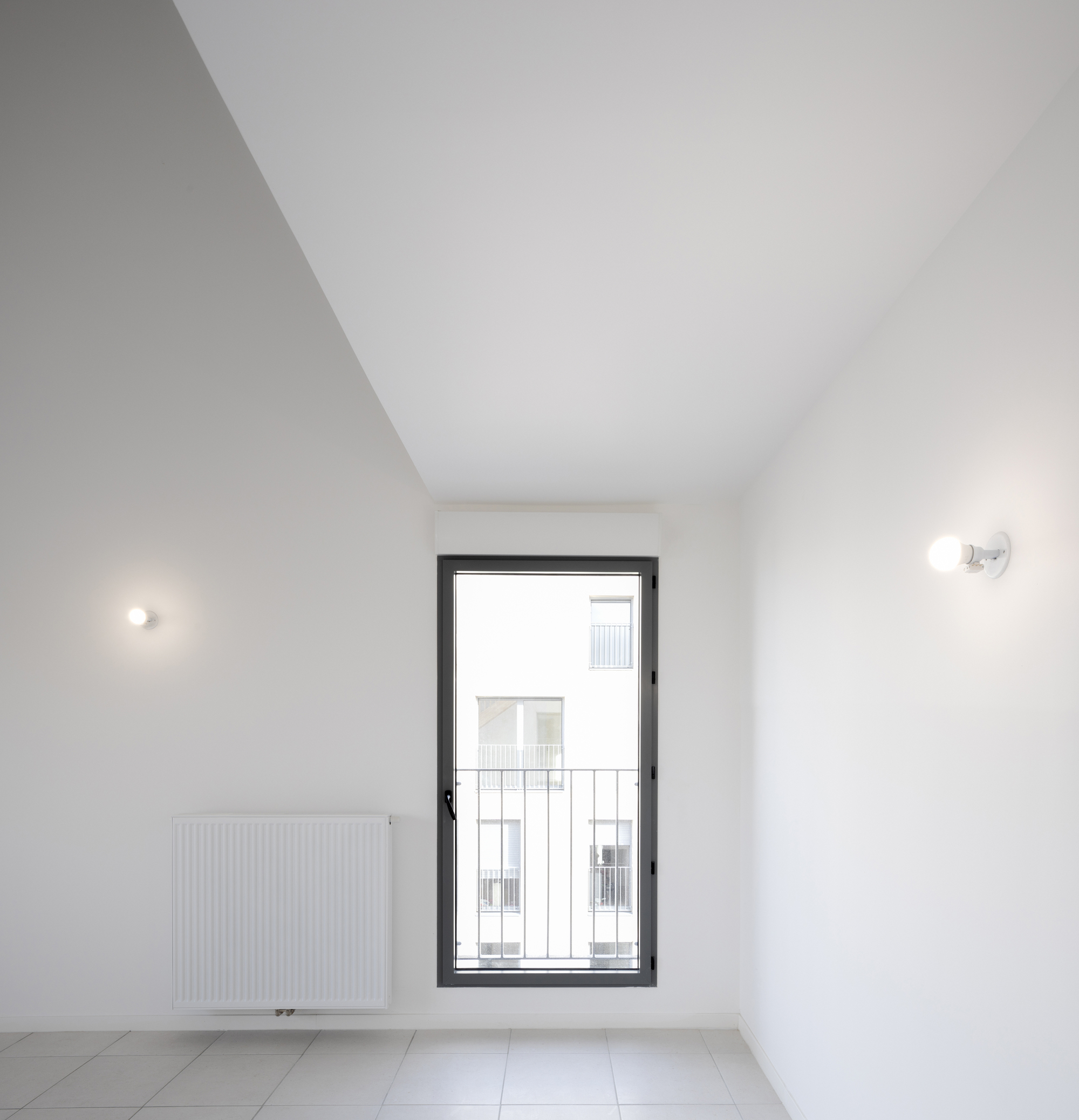 White interior room with a tall narrow window, wall-mounted sconces, and a panel radiator