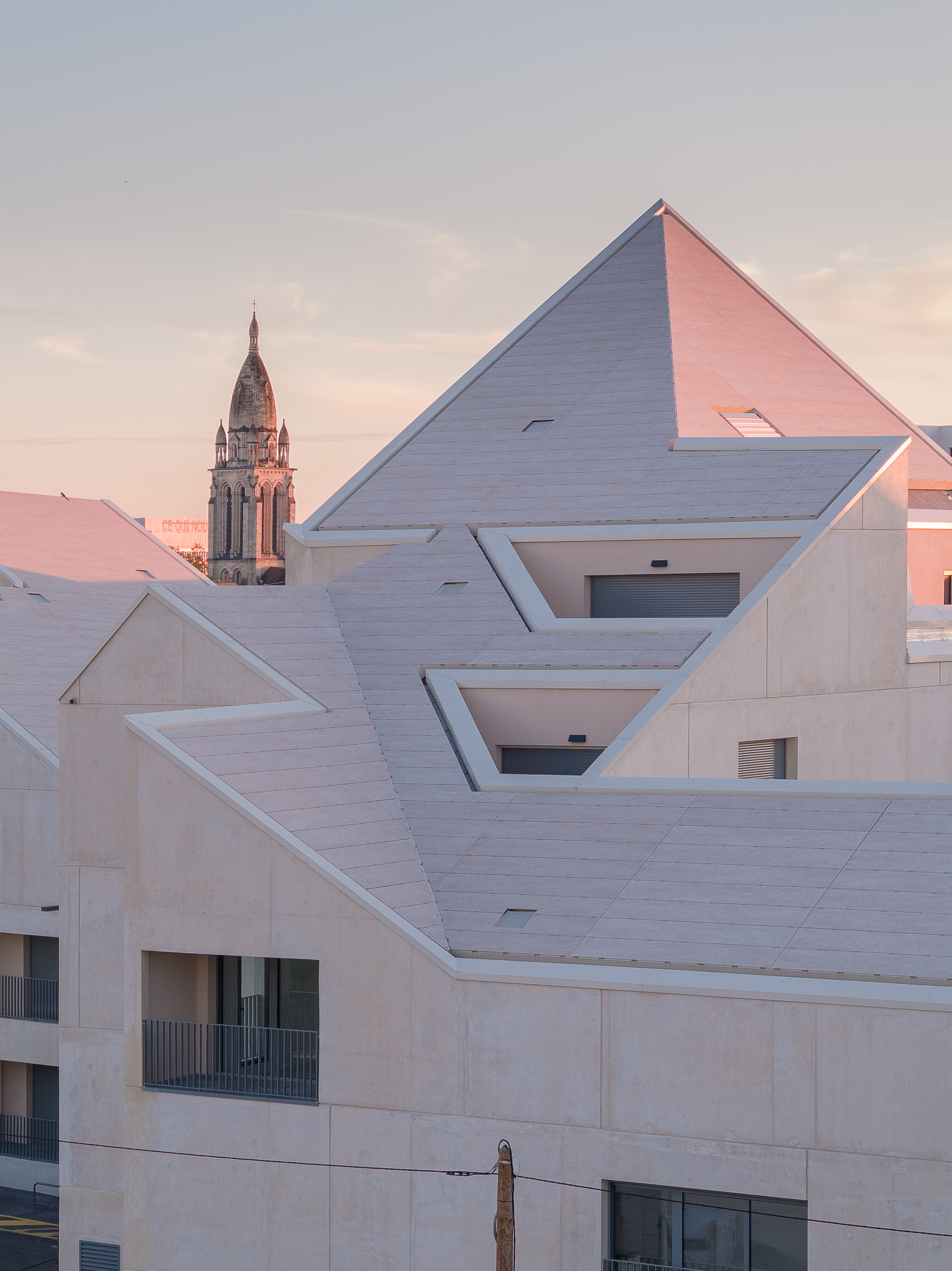 Stepped roofline with triangular gables and recessed balconies at dusk with a church tower beyond