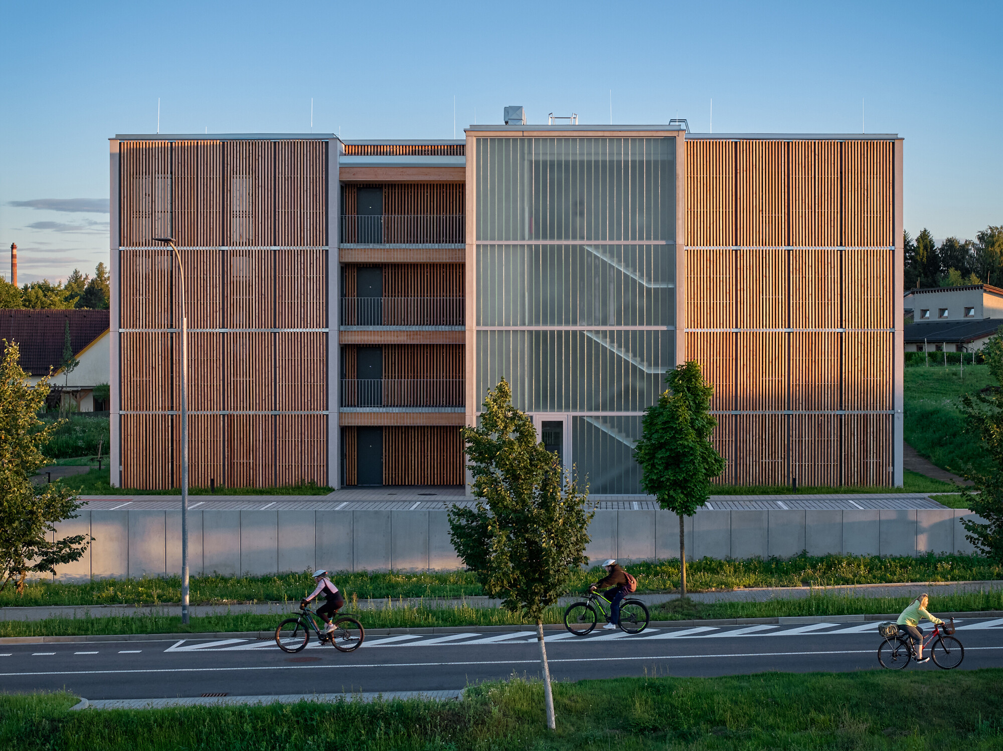 Street facade with alternating timber and translucent panel screens as three cyclists pass at dusk