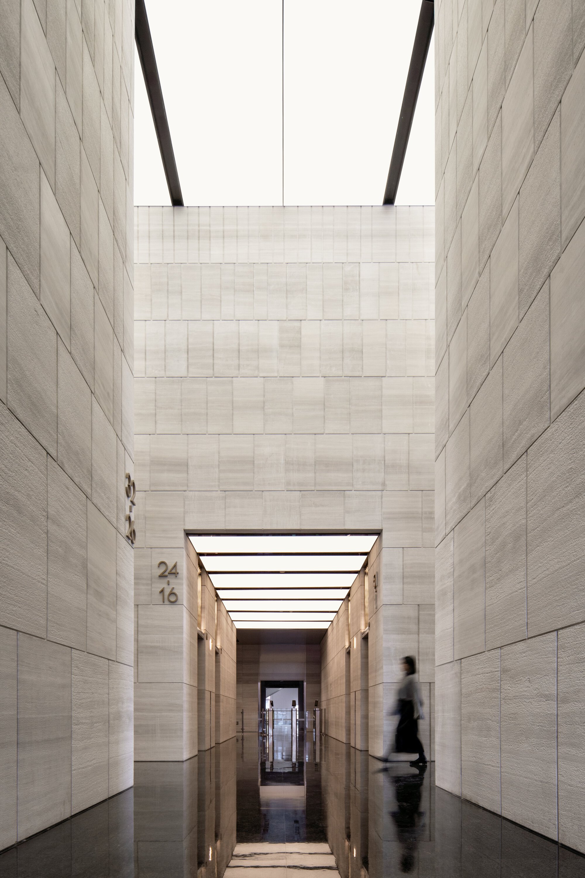 Tall atrium void lined with stone panels, a figure passing through the corridor below