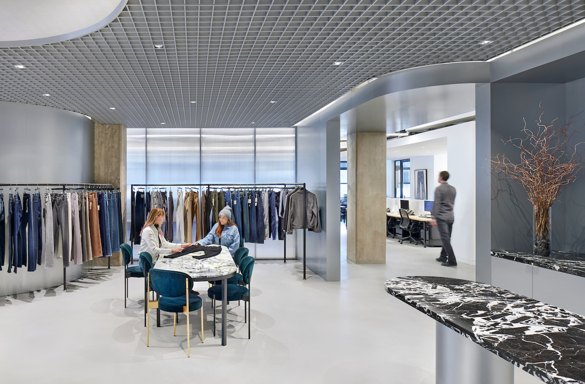 View through showroom space showing clothing displays and marbled reception desk with dried branch arrangement
