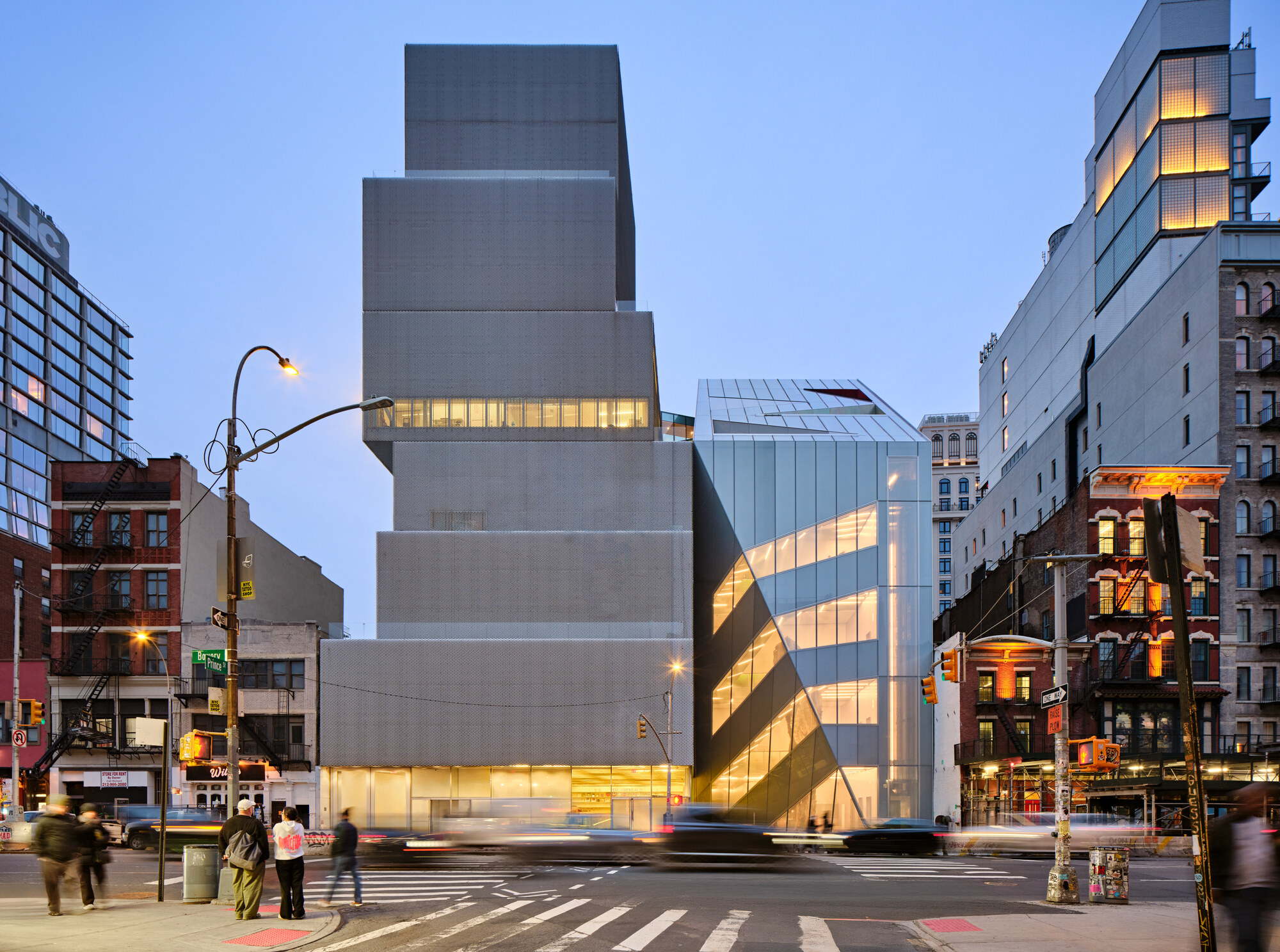 Street view of the illuminated metal facade with angled glass volumes at dusk