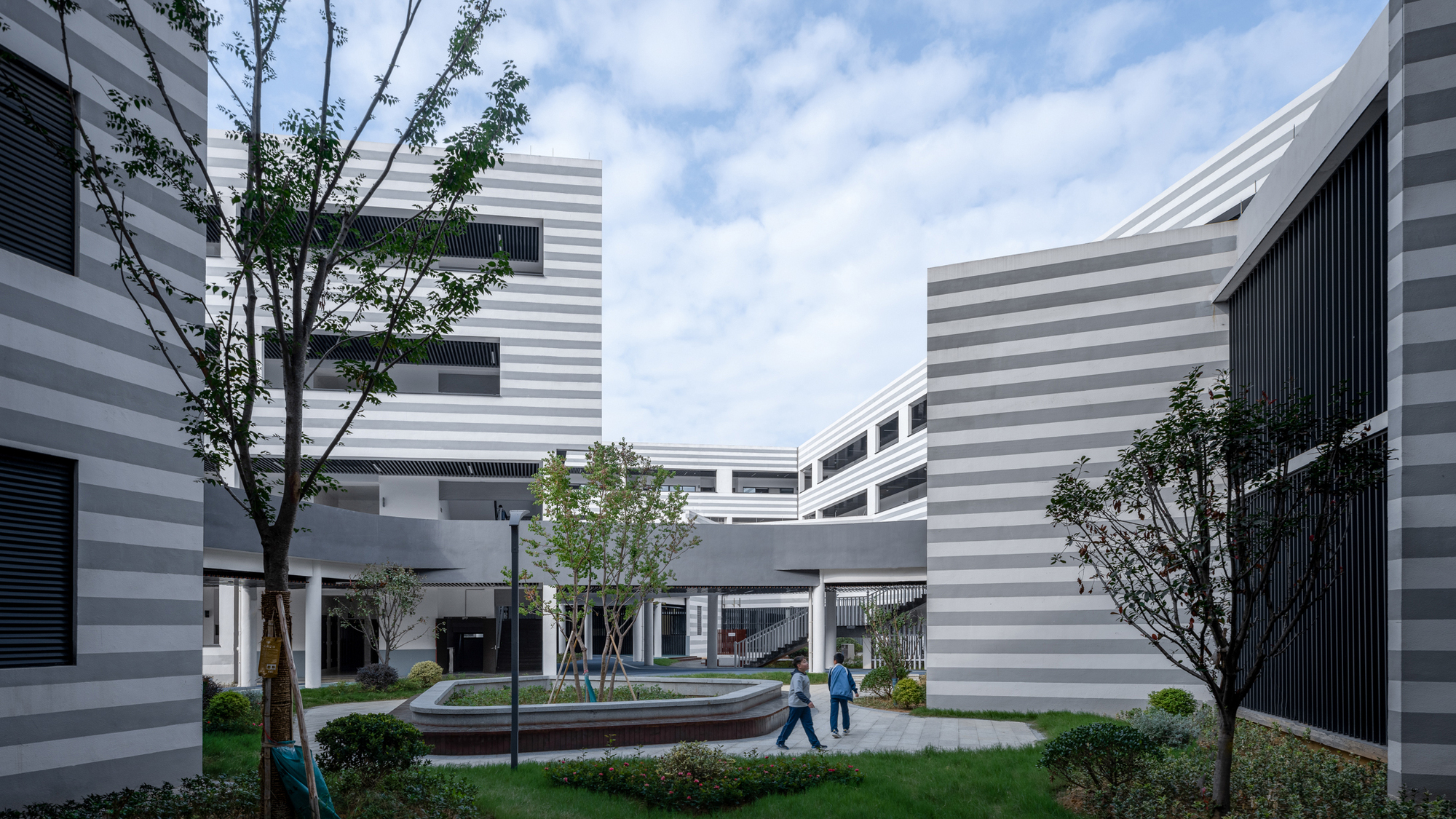 Courtyard with circular seating element and horizontal striped facades framed by trees and planted beds