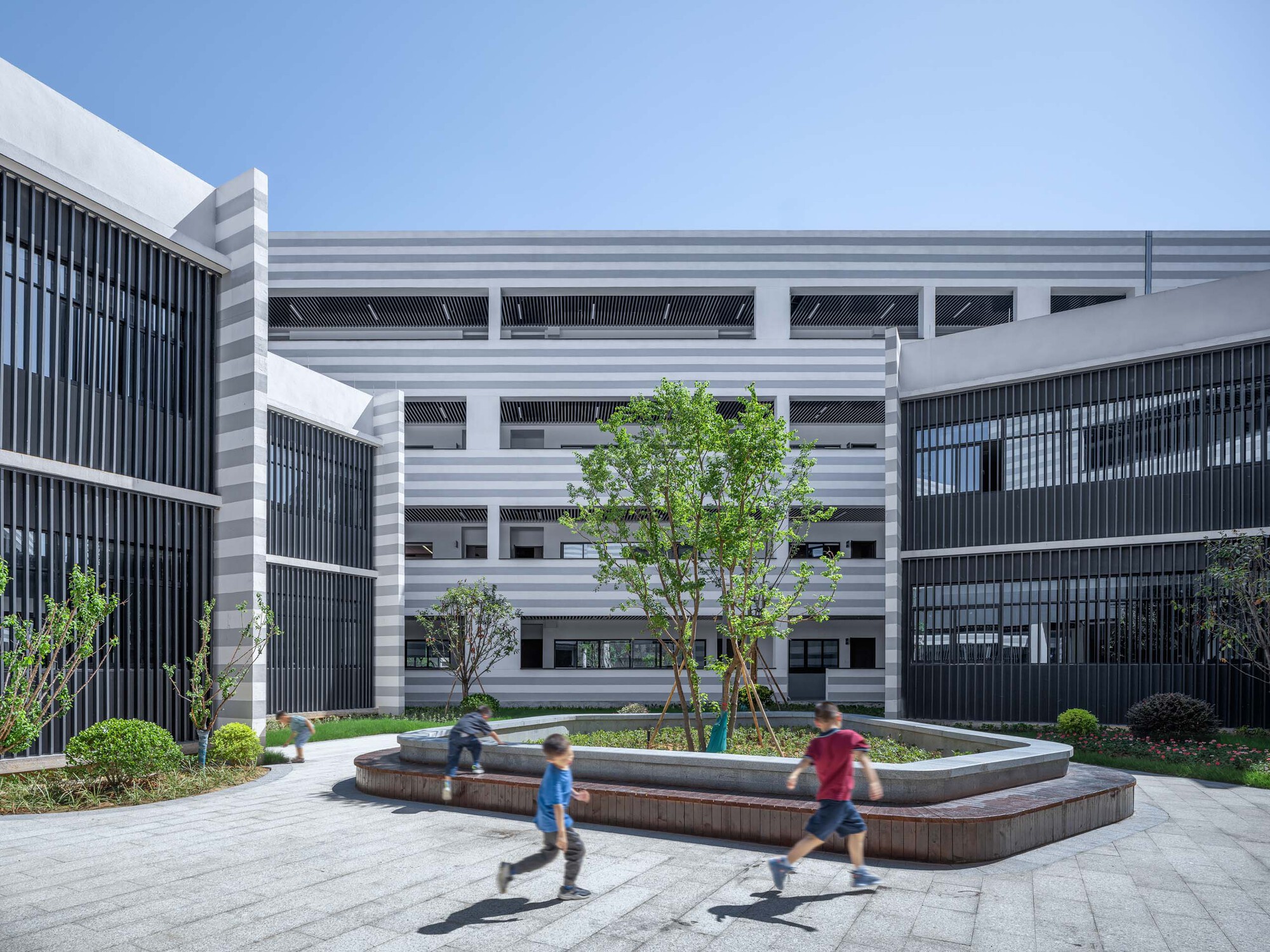 Central courtyard with circular fountain and children running past planted trees between striped facades