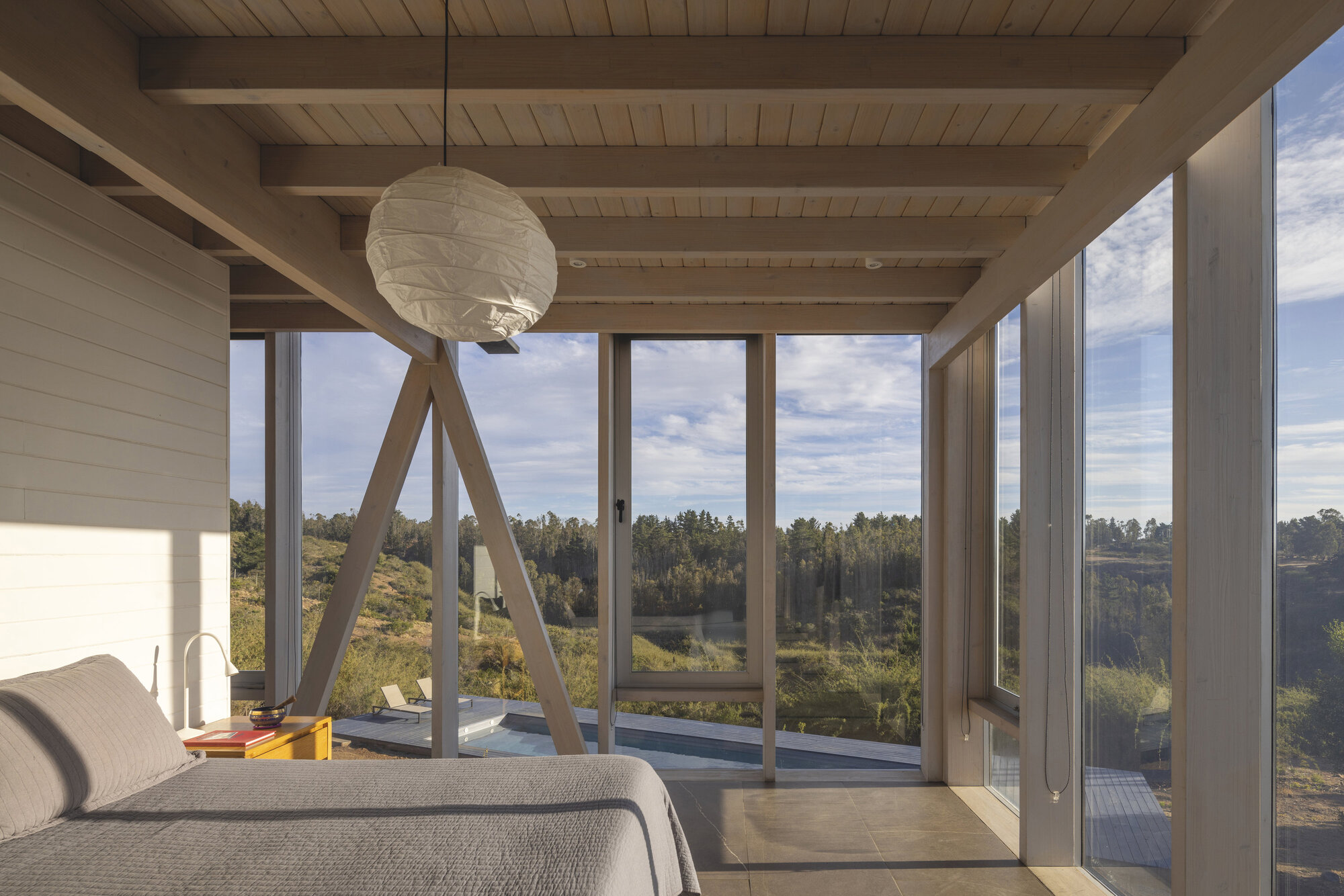 Bedroom with exposed timber beams and diagonal steel bracing framing a view of wooded hillside