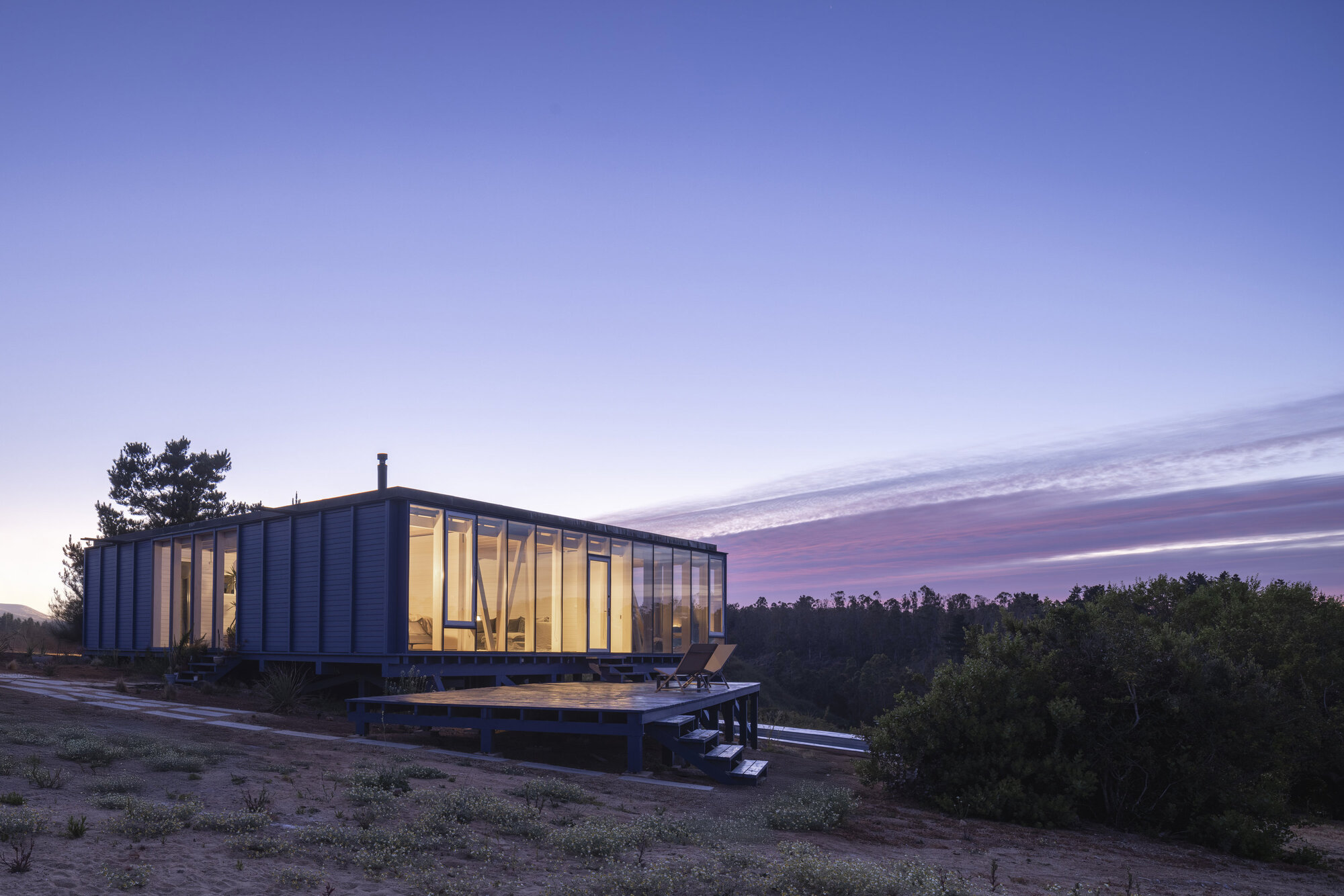 Illuminated glass-enclosed volume on raised timber platform against a sunset sky