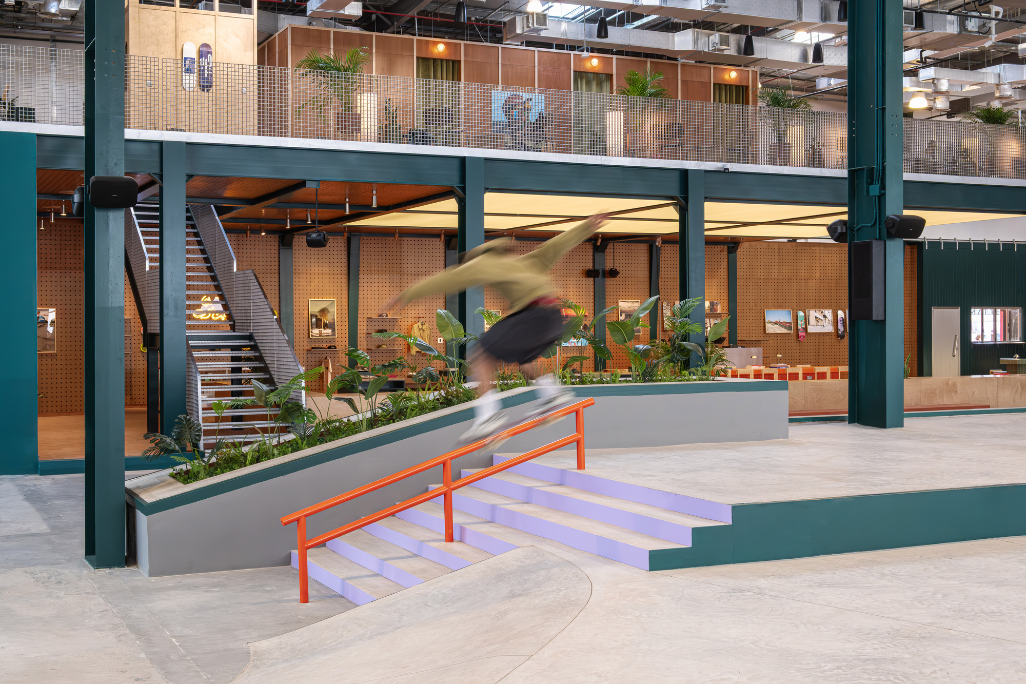 Double-height atrium with green steel columns, concrete skate ramp and a blurred skateboarder in motion