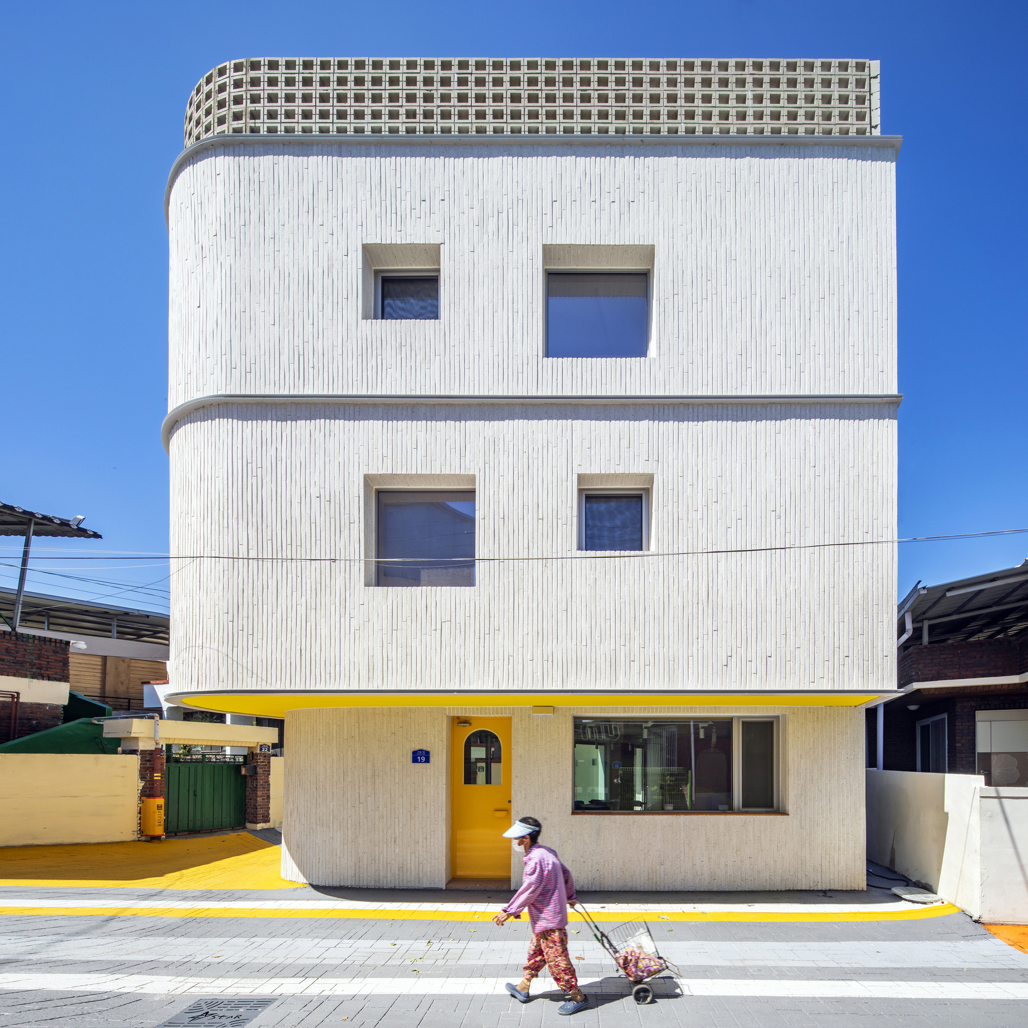 Street facade with vertical ribbed white cladding, square windows, and yellow ground floor with a person passing by