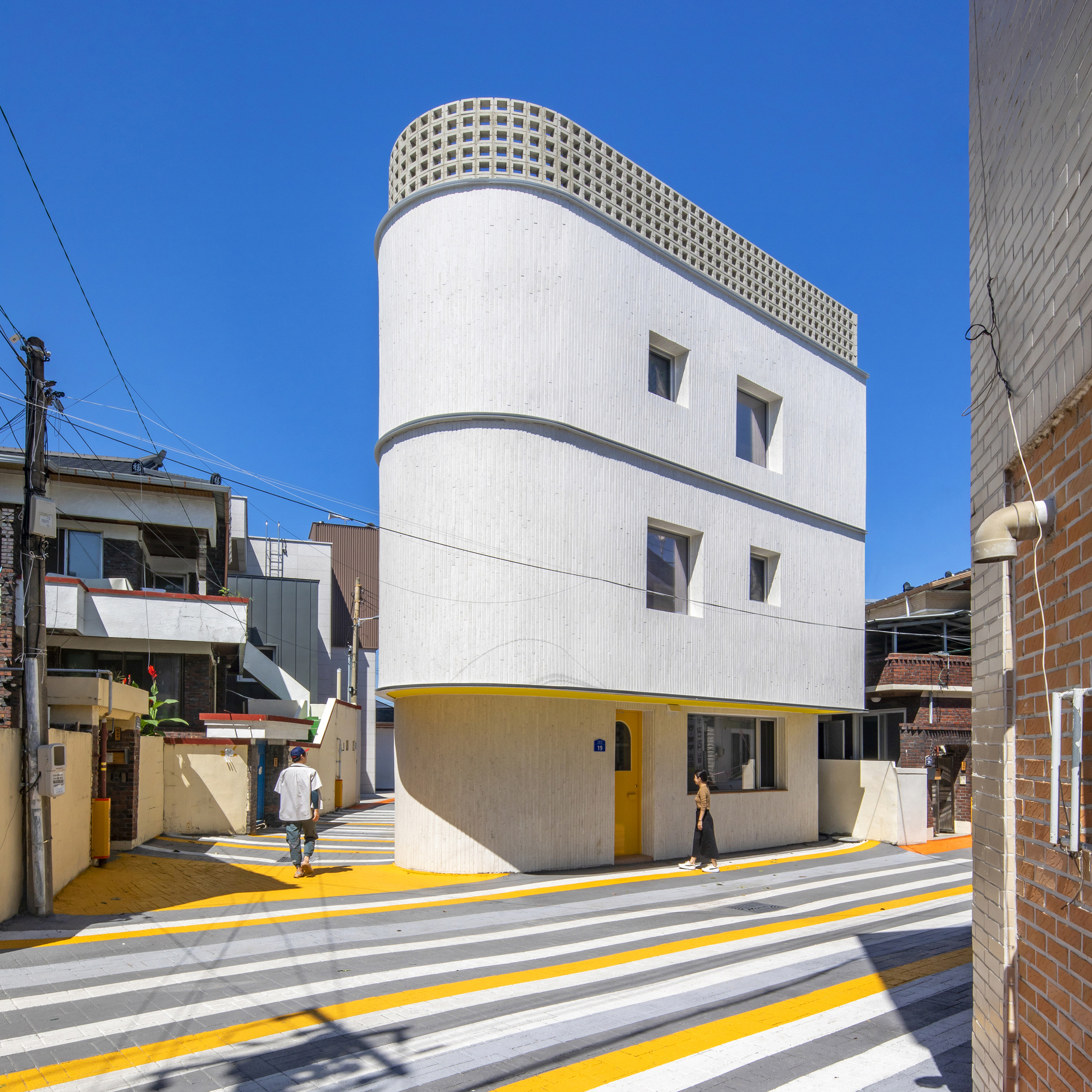 Three-story cylindrical volume with white facade, lattice rooftop parapet, and yellow-striped crosswalk extending toward the entrance