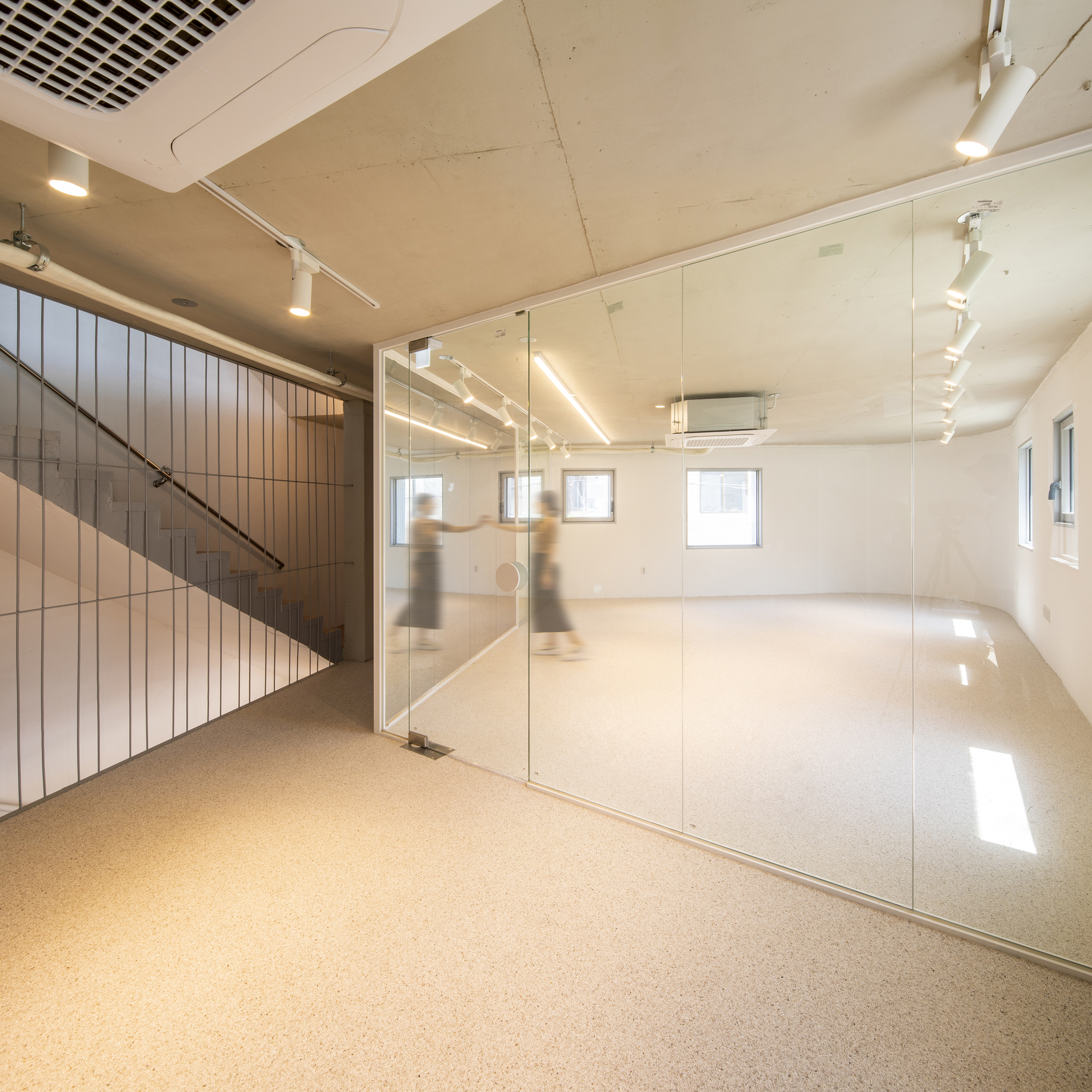 Interior space with glass partition, exposed plywood ceiling, track lighting, and blurred figures moving near the stairwell