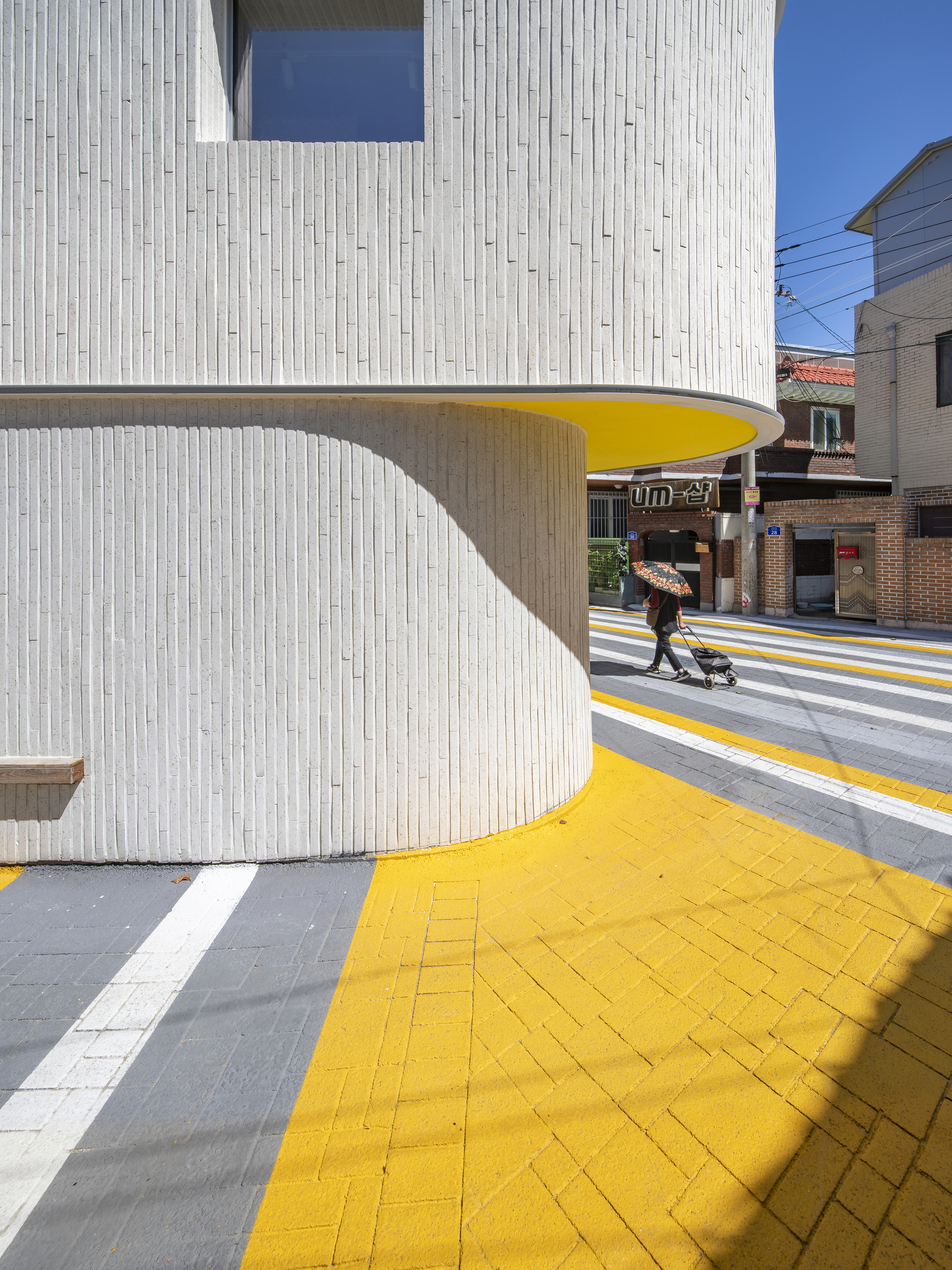 Close-up of the curving white facade underside painted bright yellow, sheltering the yellow-paved sidewalk with a pedestrian passing