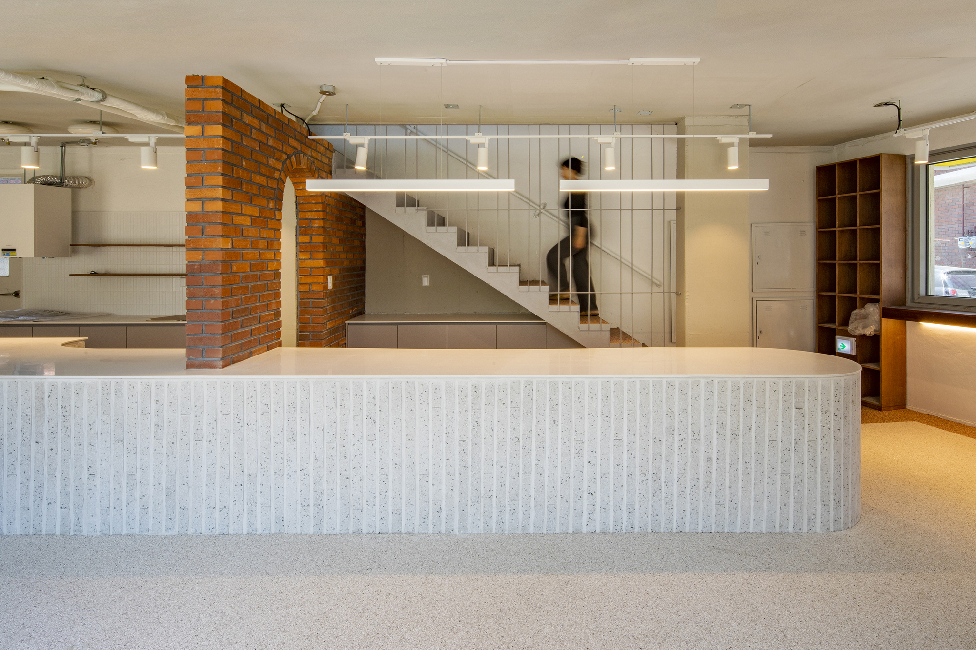 Open-plan interior with ribbed counter, glass-enclosed staircase, and exposed brick wall under fluorescent lighting