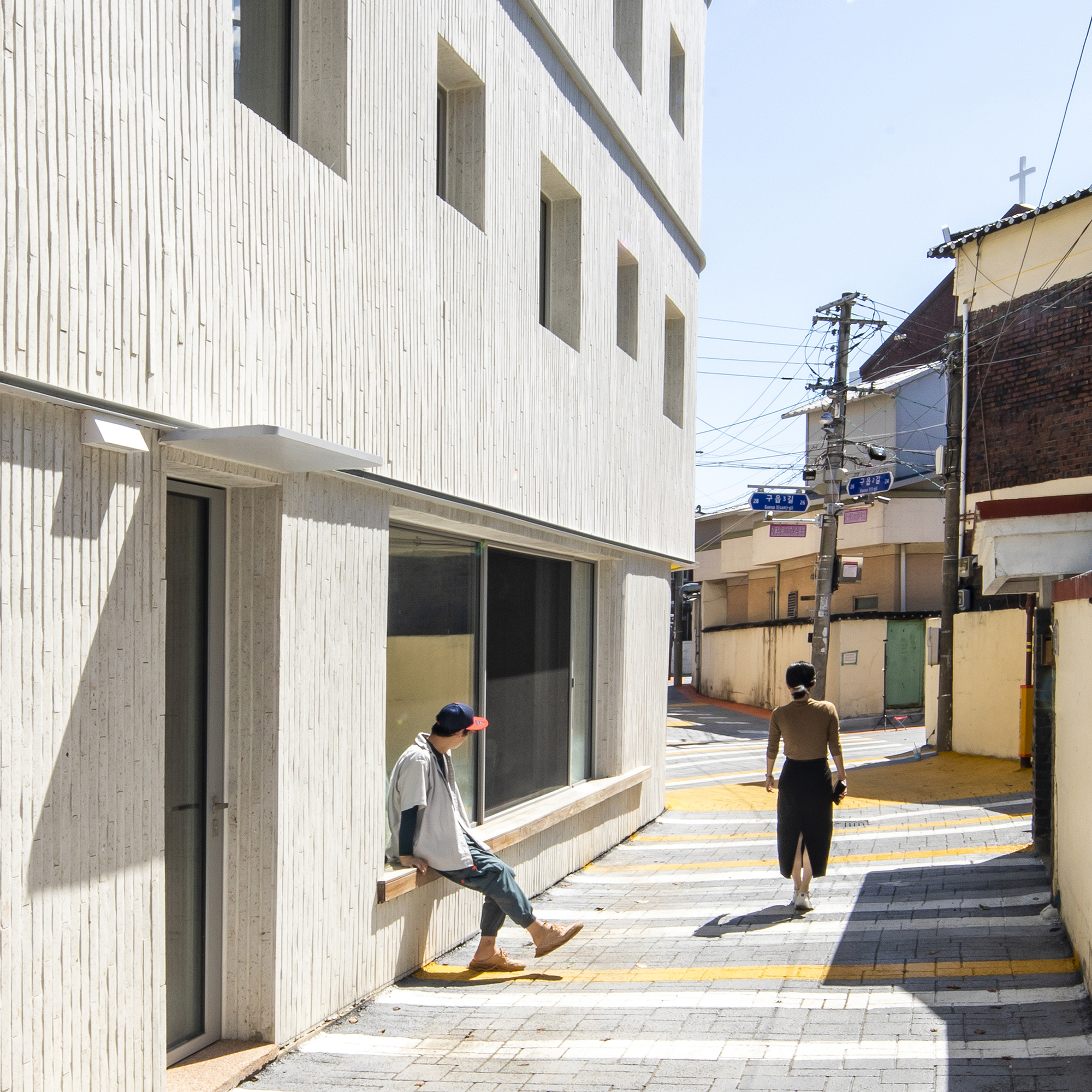 Entrance elevation with vertical cladding and yellow-striped pavement where two figures walk in morning light