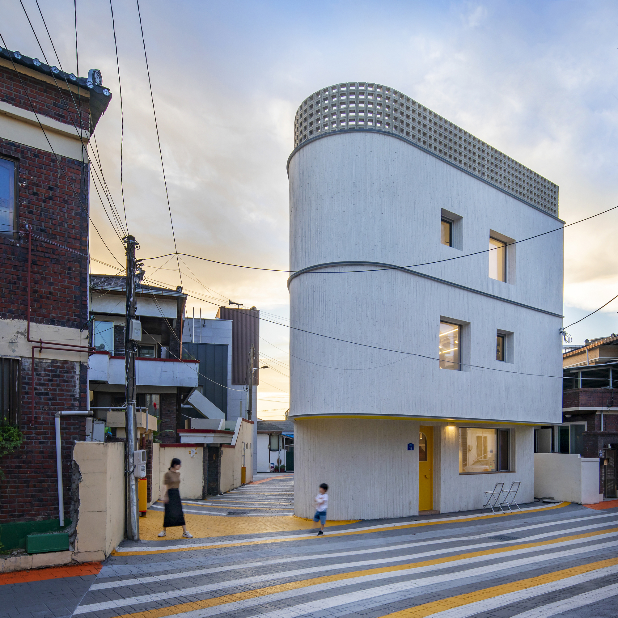 Curved white facade with perforated parapet at the corner of a striped pedestrian crosswalk at dusk