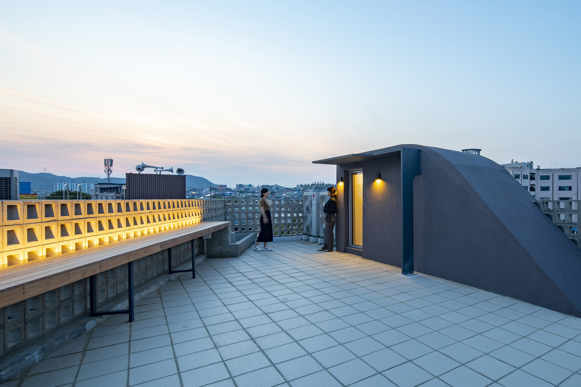 Rooftop terrace with tiled paving and illuminated perforated screen wall overlooking the neighborhood at dusk