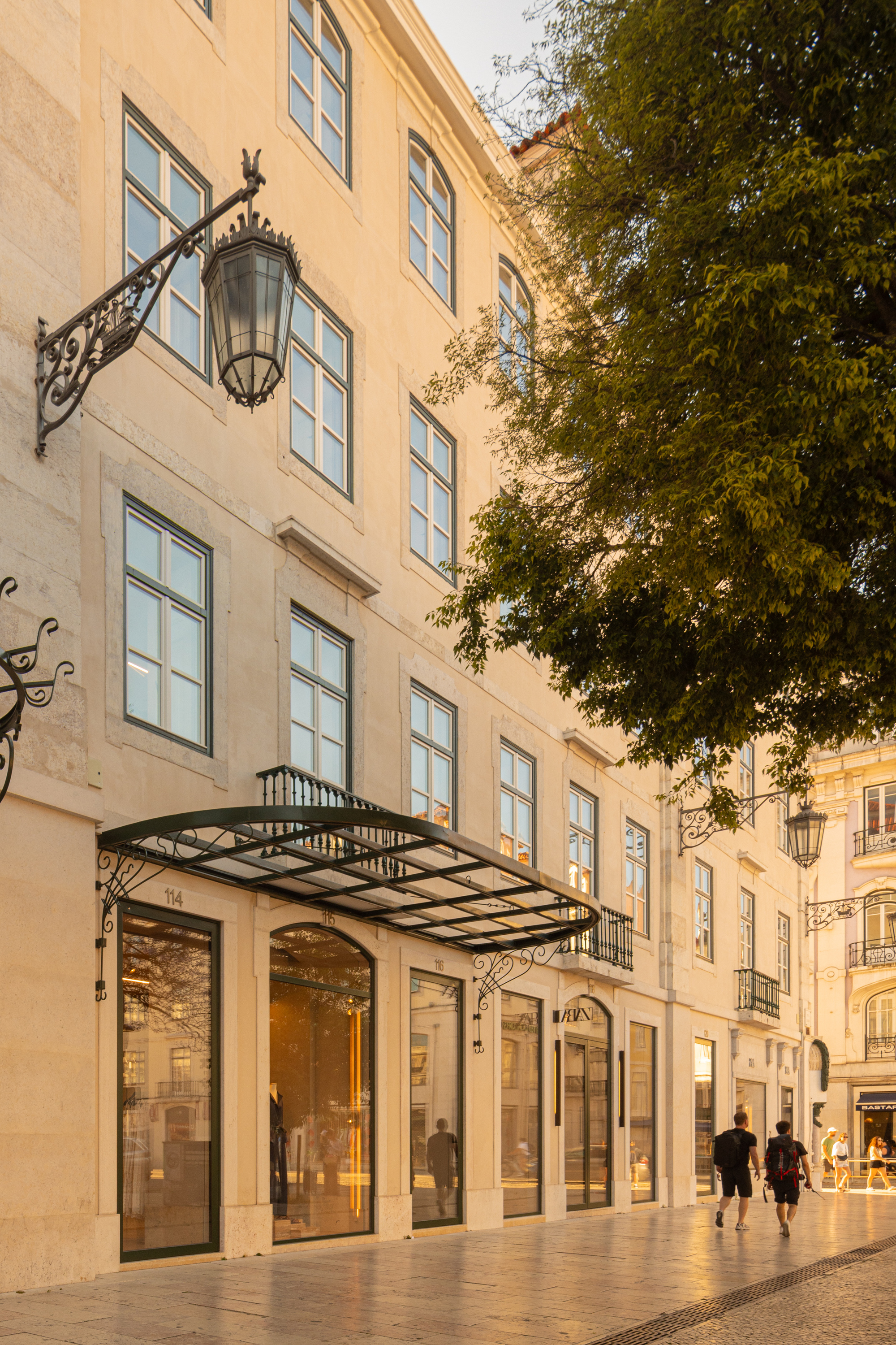 Street facade with wrought iron canopy sheltering glazed storefronts as pedestrians pass at twilight