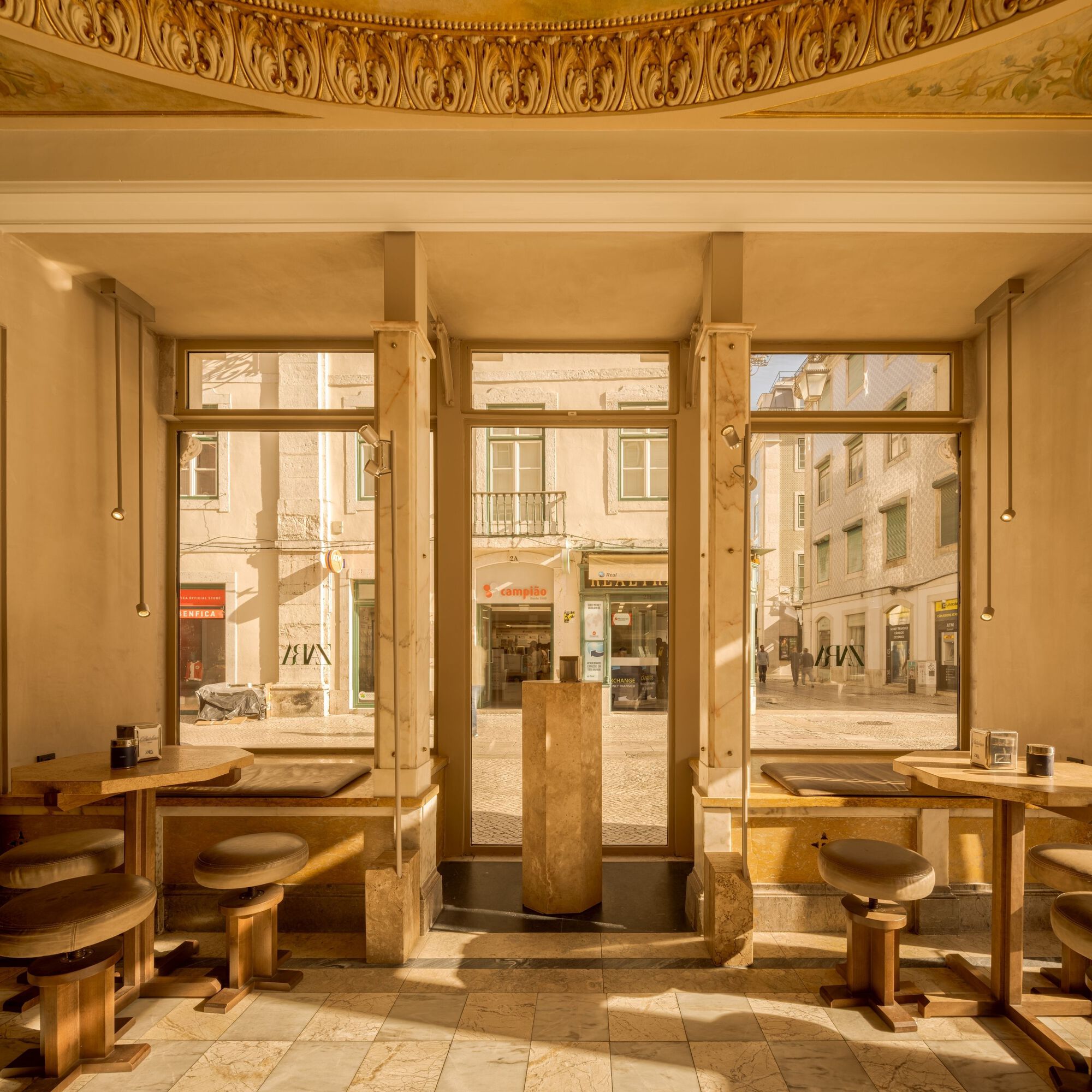 Interior seating area with tall windows framing the street and timber tables beneath a frescoed ceiling